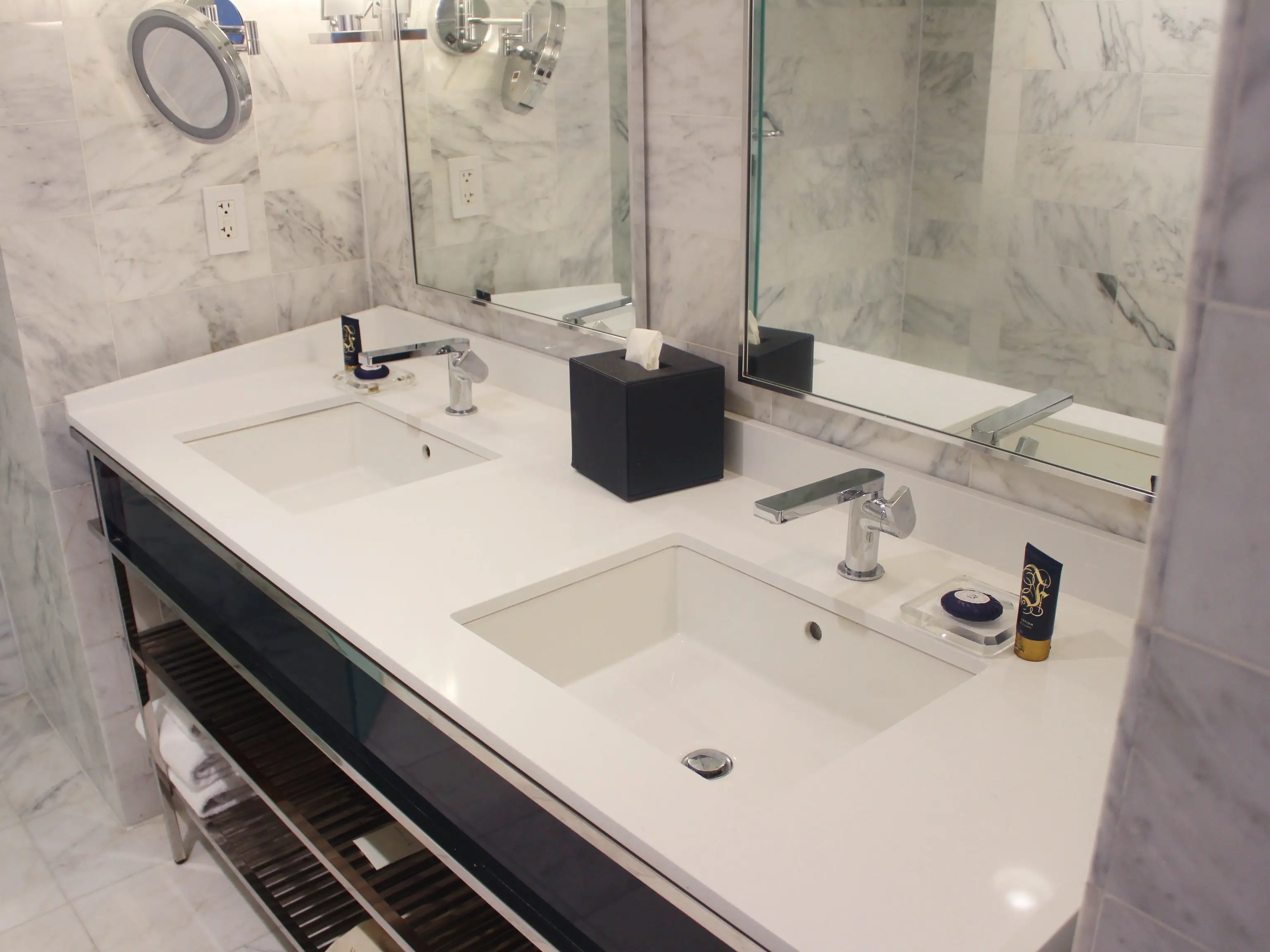 Double-sink bathroom vanity with white countertops, marble walls, mirrors, faucets, toiletries, and a black tissue box.
