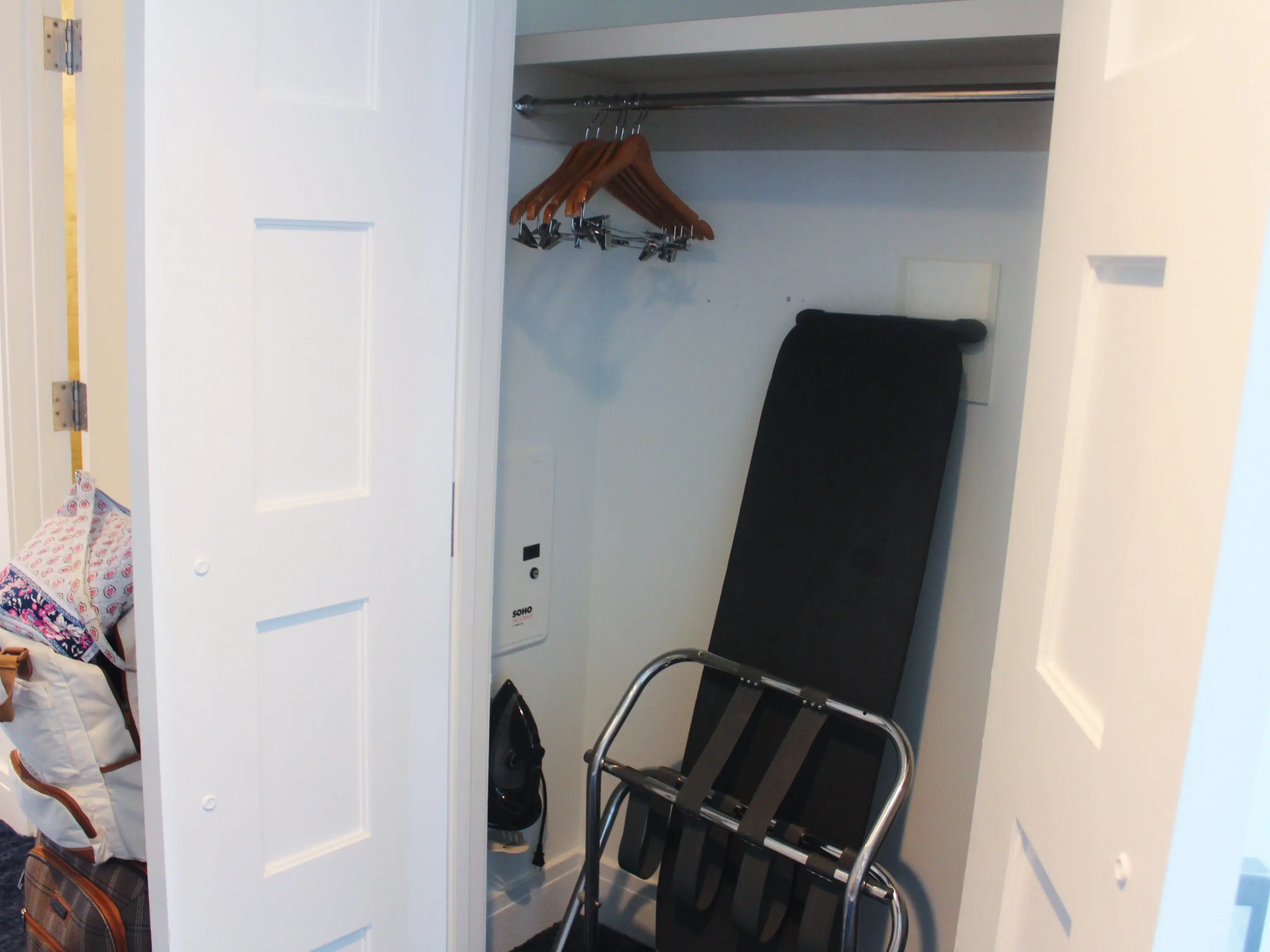 Open hotel closet with wooden hangers, a black luggage rack, bags, and white paneled doors.