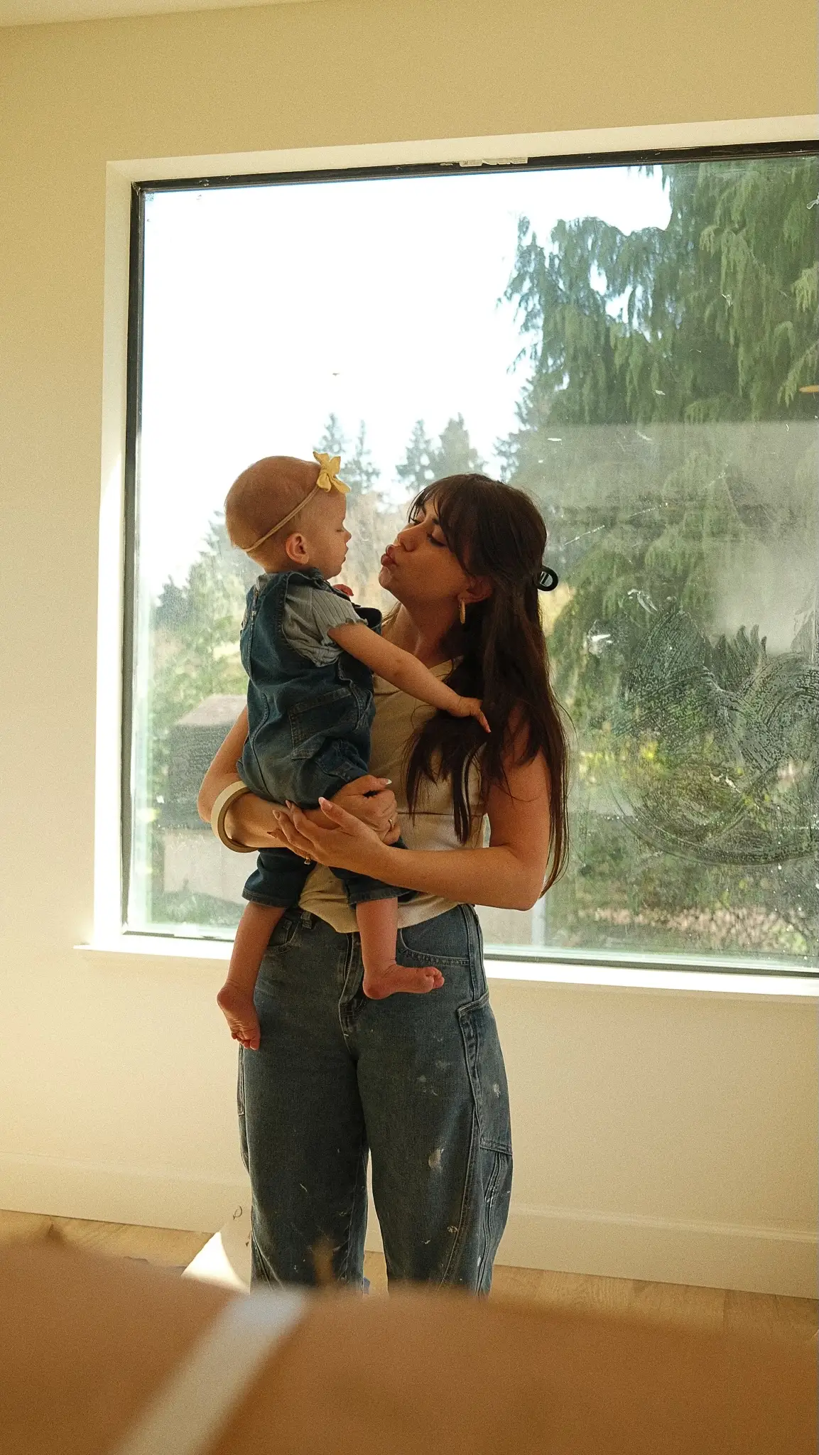 A woman holds her baby in front of a large window.