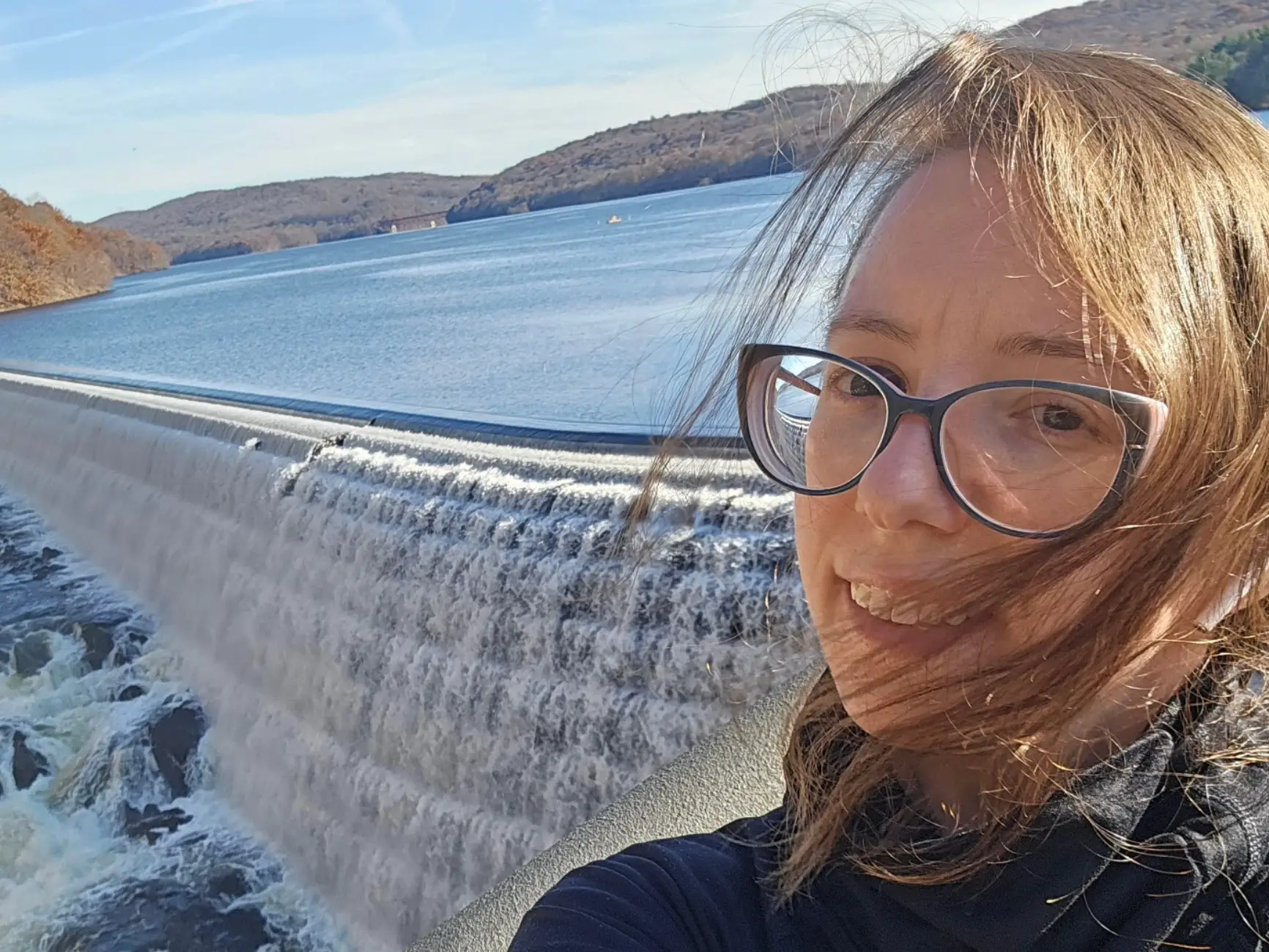 The writer standing in front of a dam in her new town, with water and hills in the background.