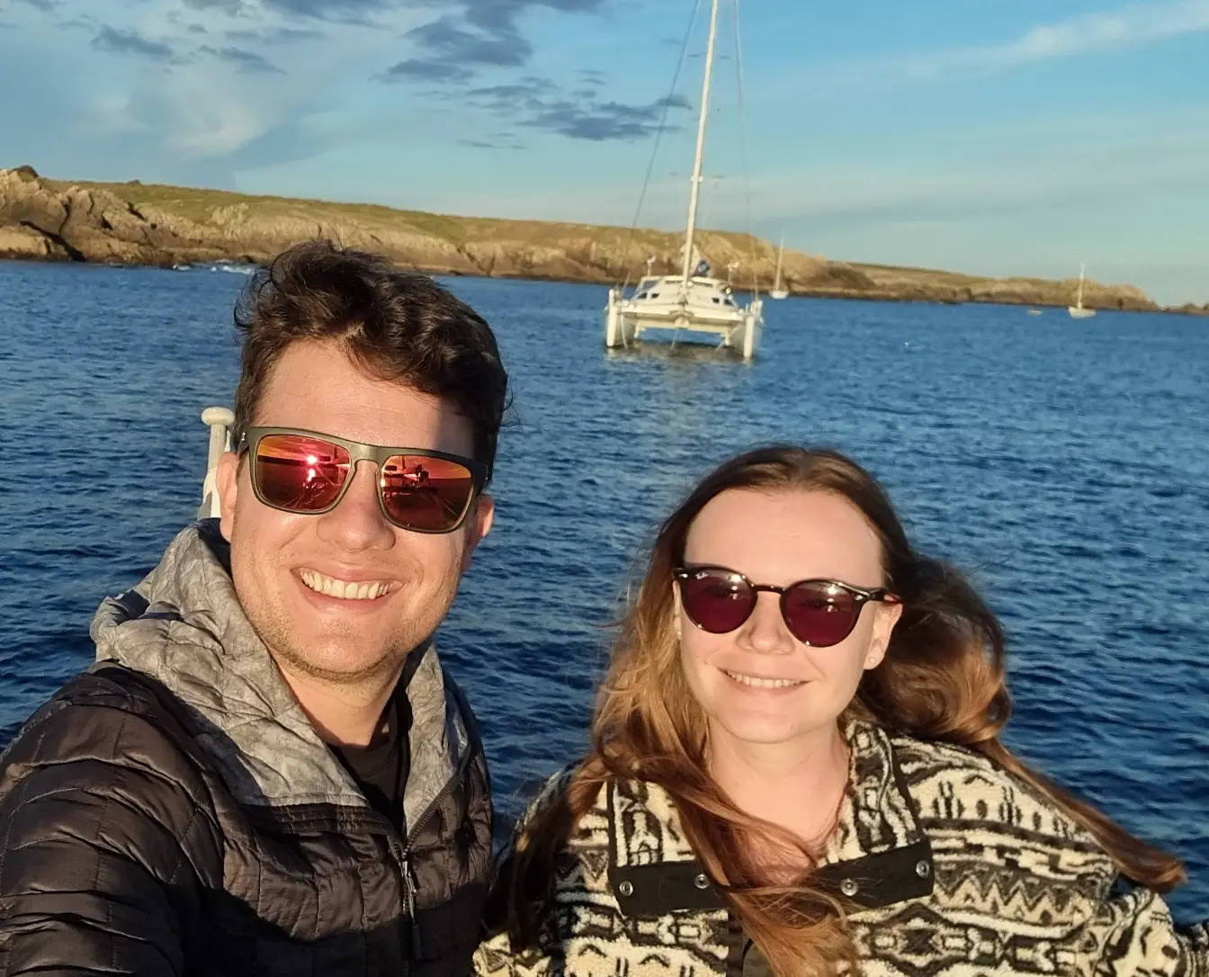 The writer and her husband standing on their boat, in front of another boat, in the blue ocean.