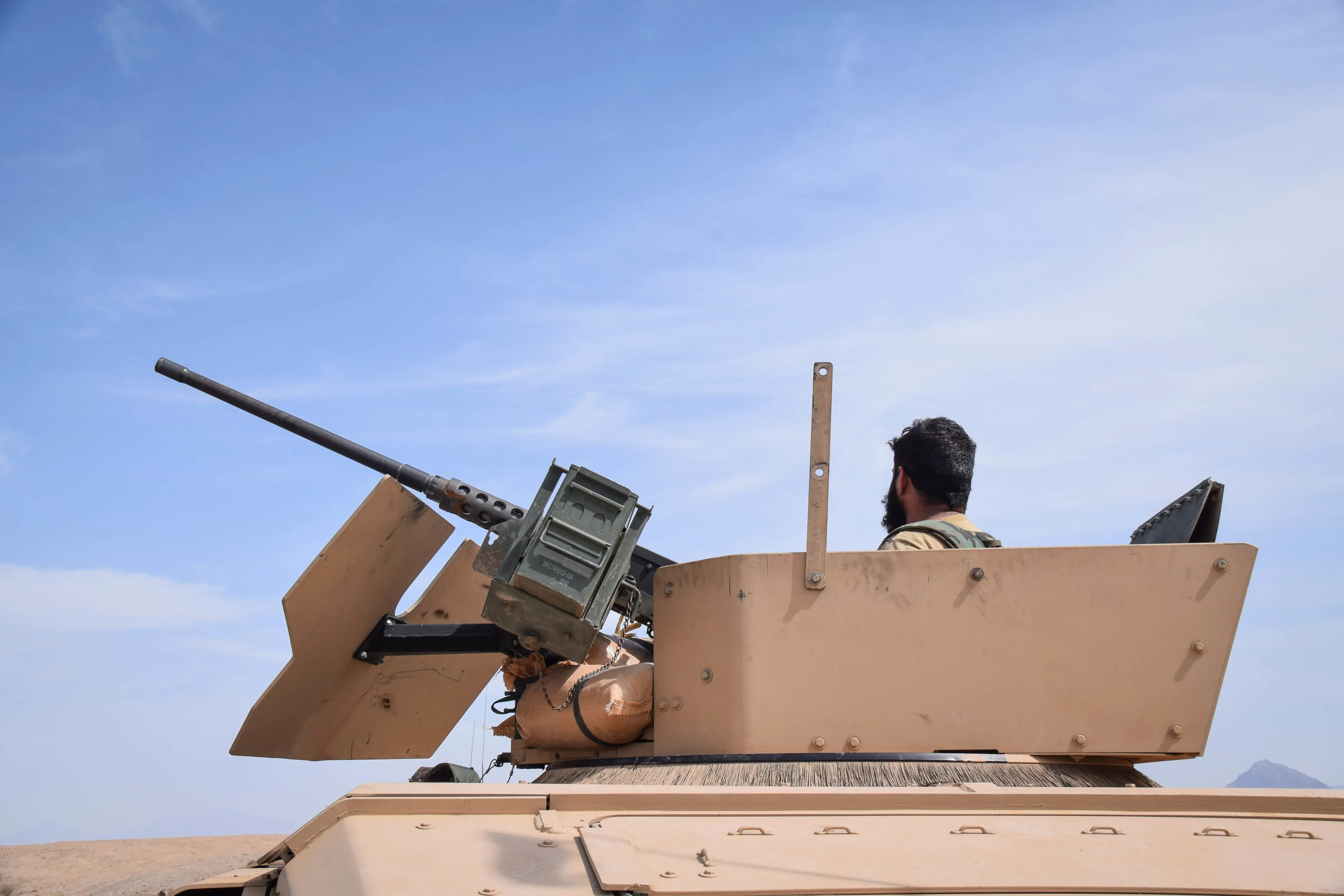 The top of a pale colored armored vehicle against a blue sky with a large machine gun mounted on front with a man's head also visable above.