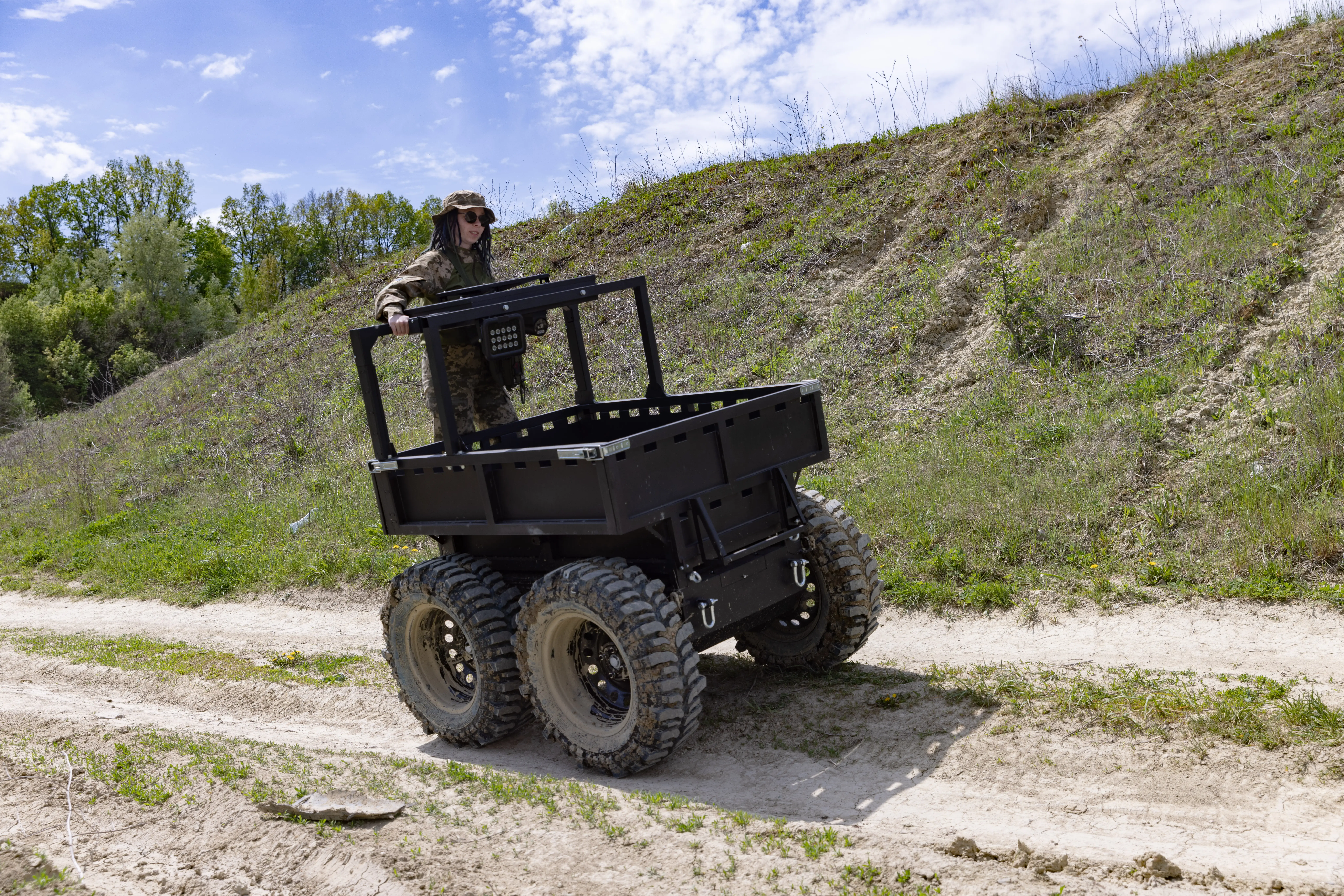 A black robot on four wheels drives on a dirt track beside grass and under a blue sky with a person in khaki inside it