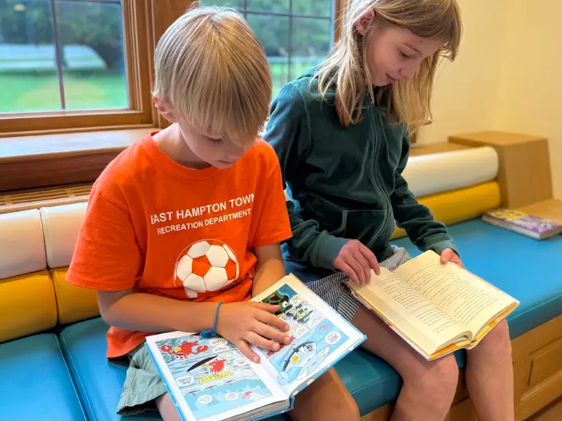 Two kids sit on a colorful bench reading books beside large windows.