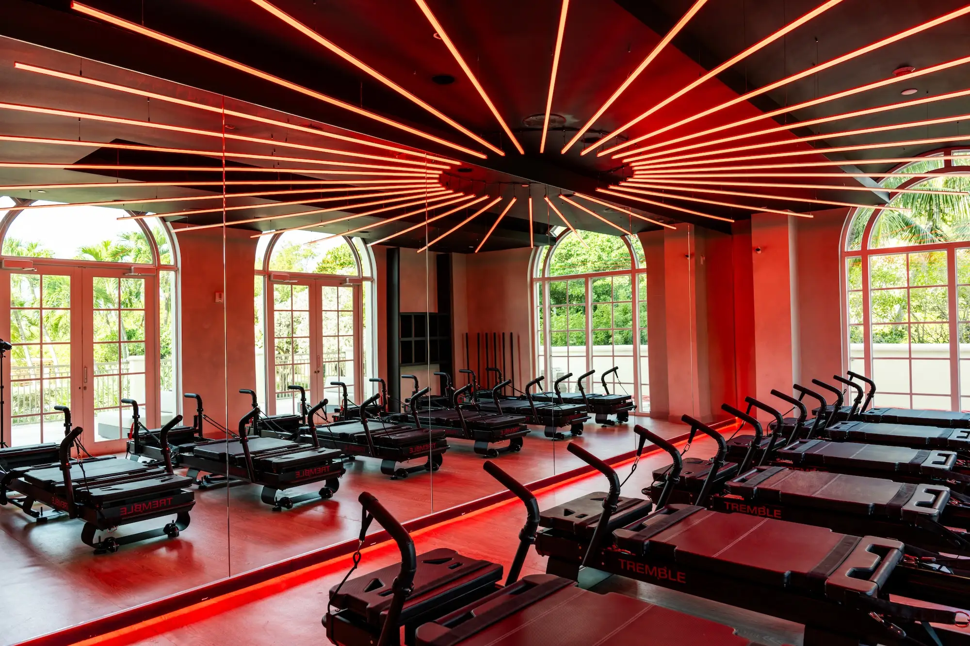 Fitness studio lined with black treadmills beneath red radial ceiling lights and arched windows.