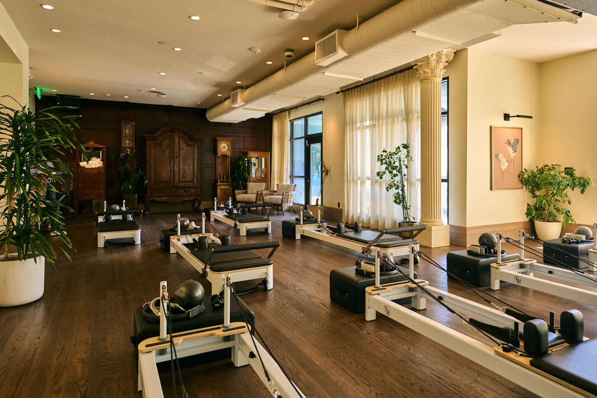 Sunlit Pilates studio with reformer machines, wood flooring, indoor plants, columns, and antique furnishings.