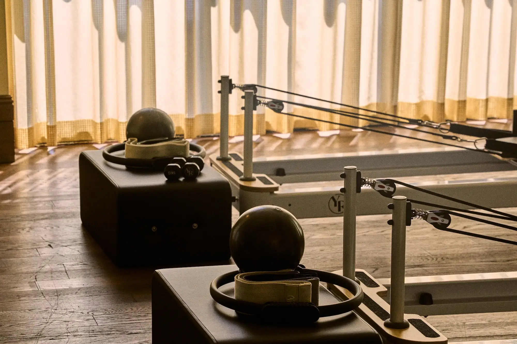Pilates reformer machines with straps, exercise balls, and head loops sit on a wooden floor beside sheer curtains.