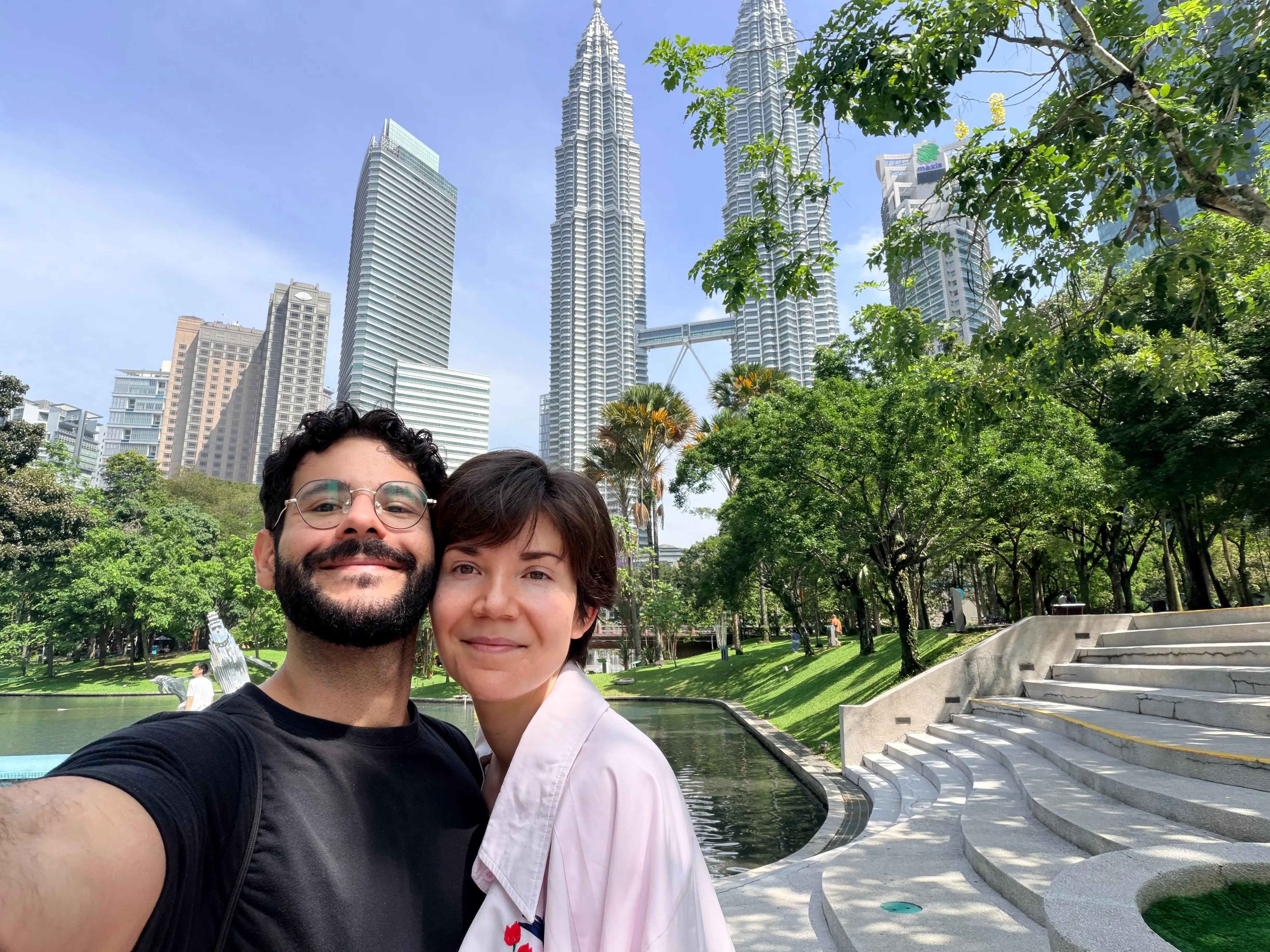 Two visitors take a selfie in a landscaped park with the Petronas Twin Towers skyline in Malaysia behind them.