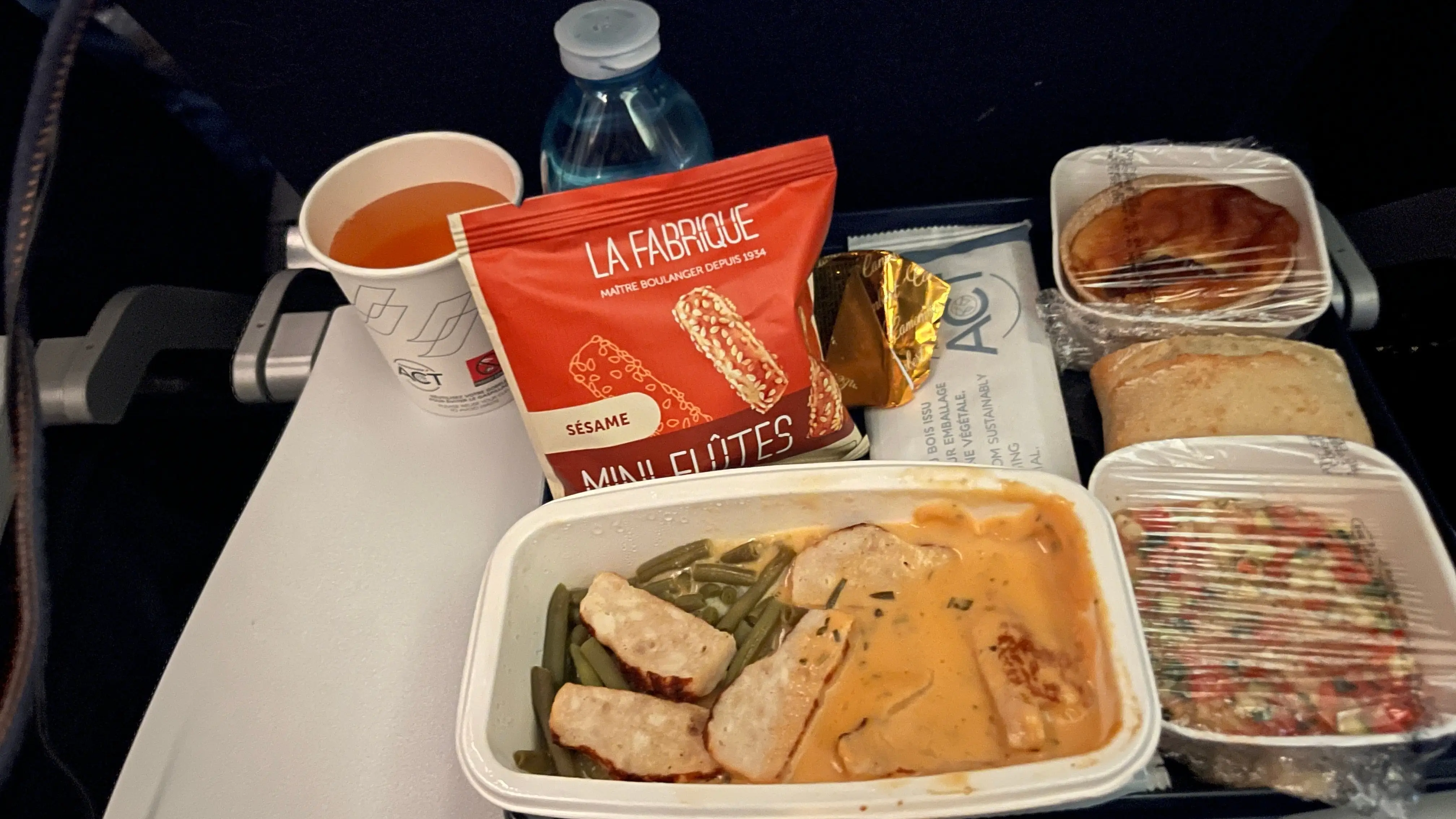 Airplane tray table with a packaged meal, bread, snacks, water bottle, and drink cup in a passenger seat.