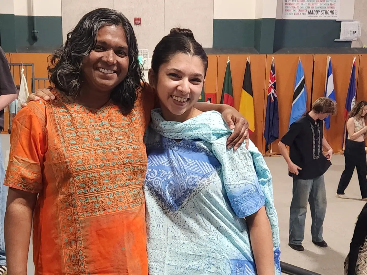 Two people wearing colorful outfits stand together on gymnasium bleachers with flags and a banner in the background.