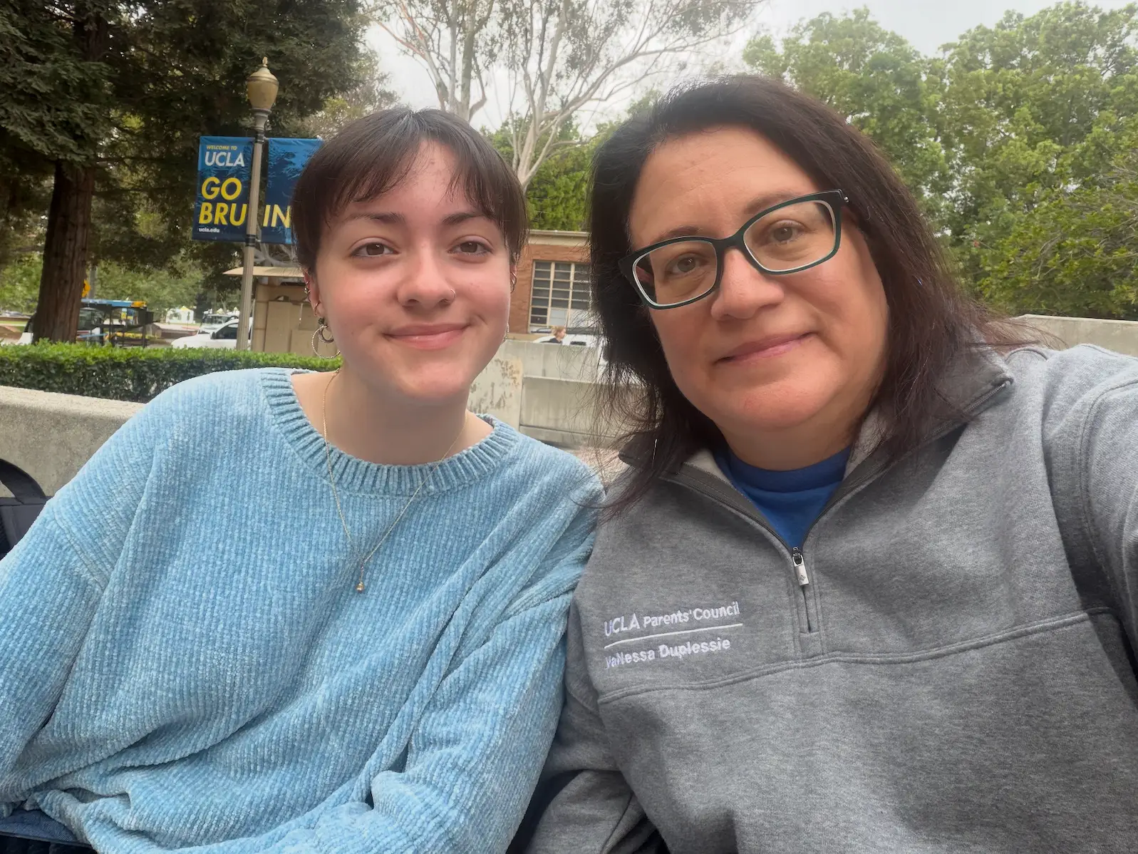 Two people sit together outdoors on a UCLA campus walkway with trees and a “GO BRUIN” banner behind them.