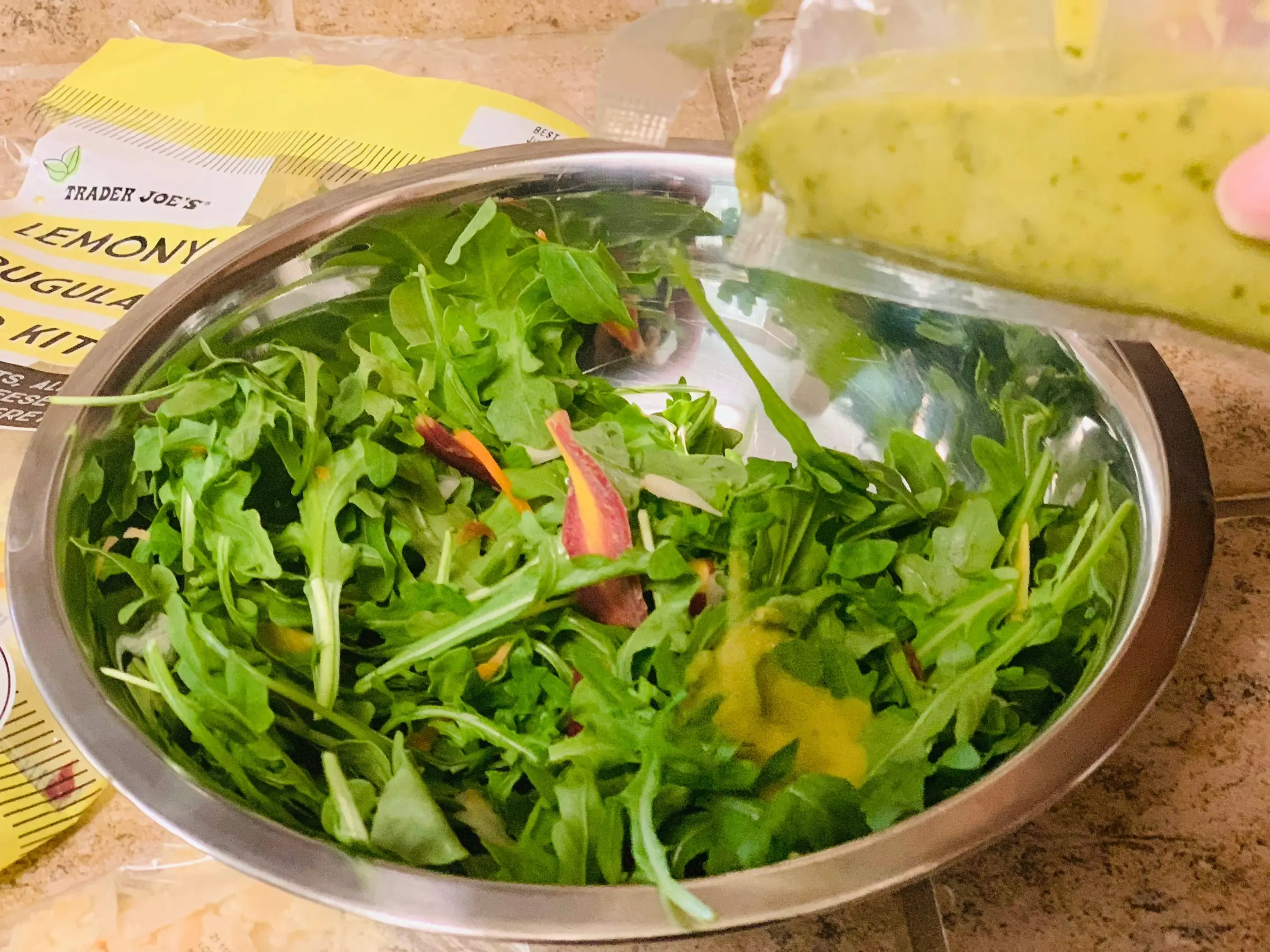 Dressing being poured on Trader Joe's lemony arugula salad, which is in a metal bowl.