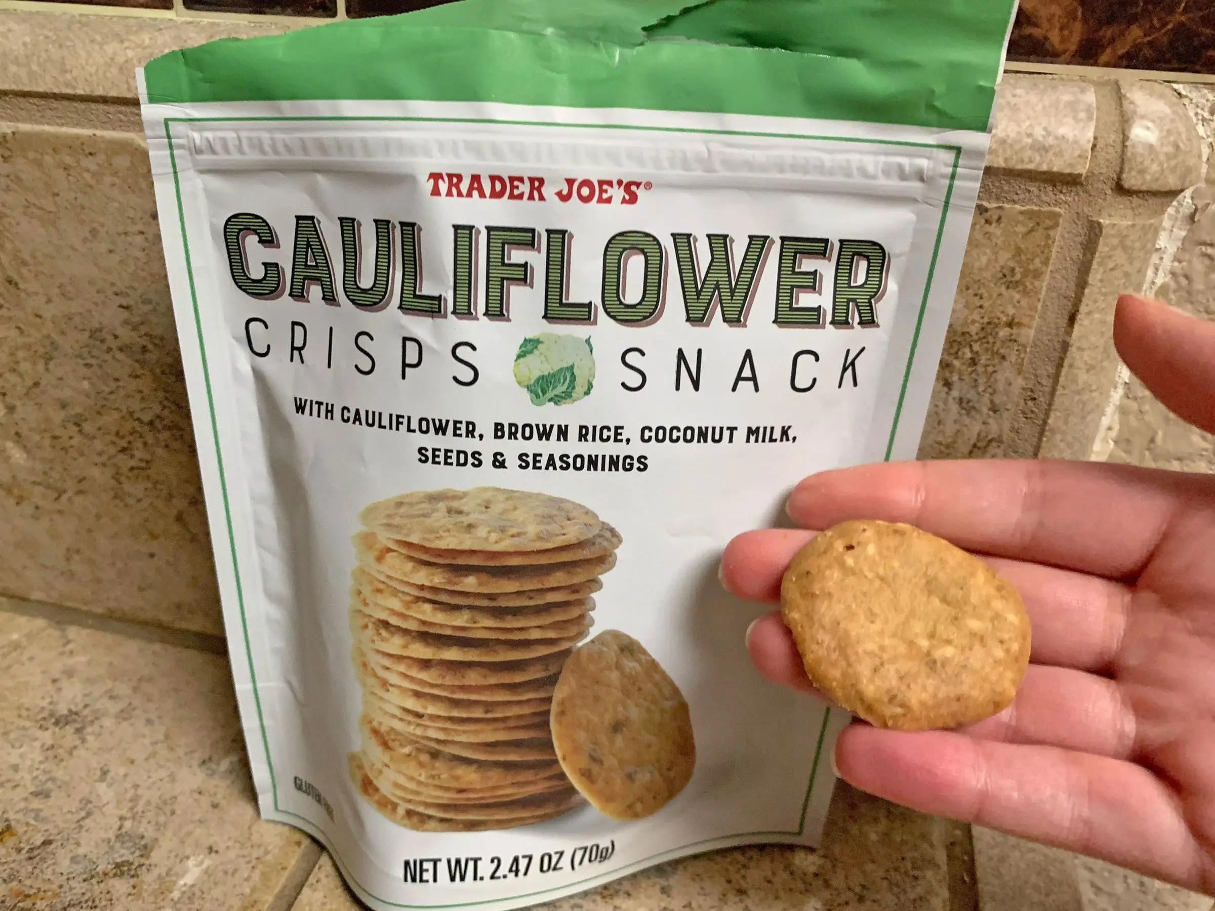 A hand holding a Trader Joe's cauliflower crisp with the bag the product came in in the background.
