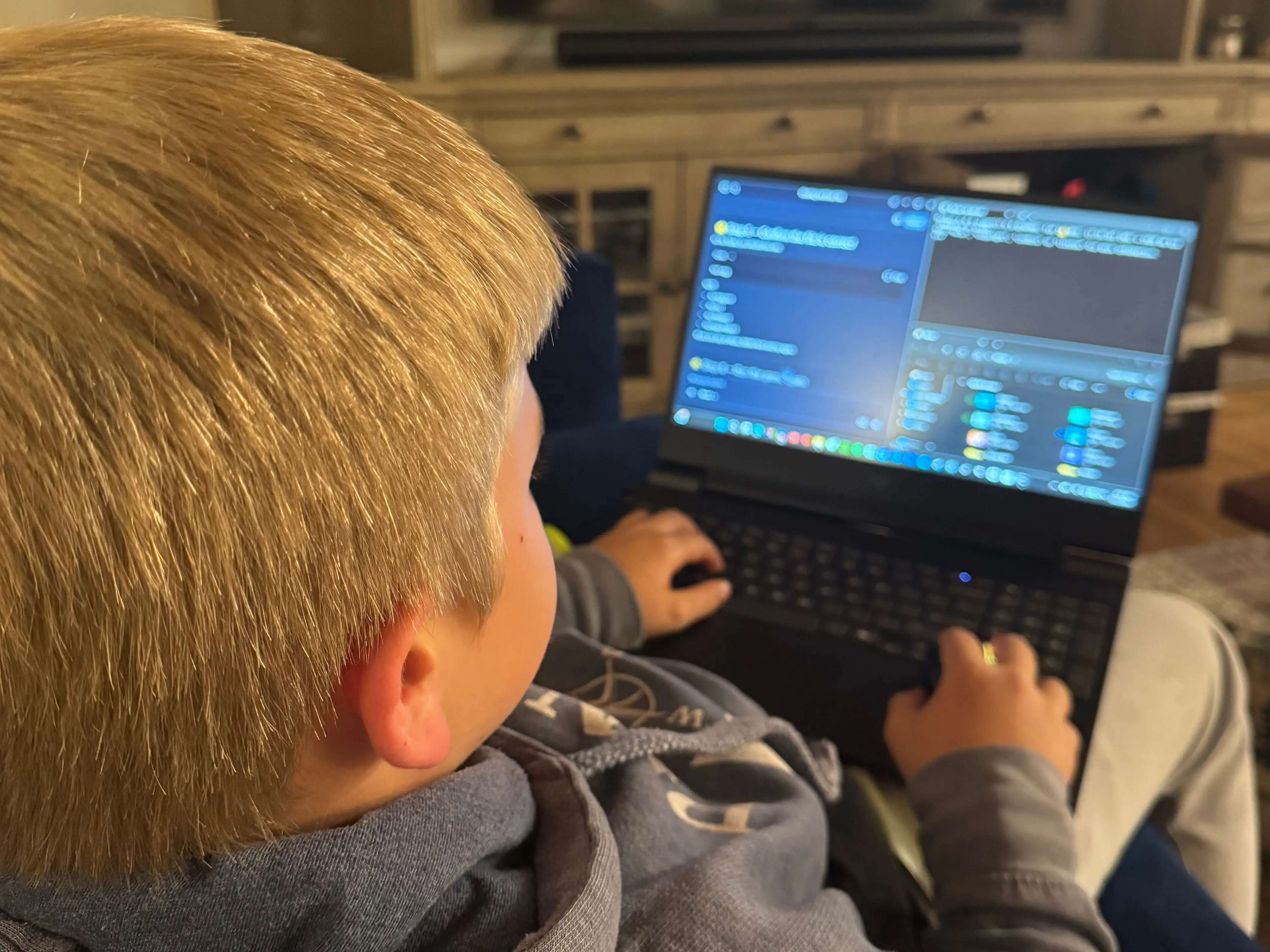 Child in a hoodie uses a laptop on a couch in a living room with a TV.
