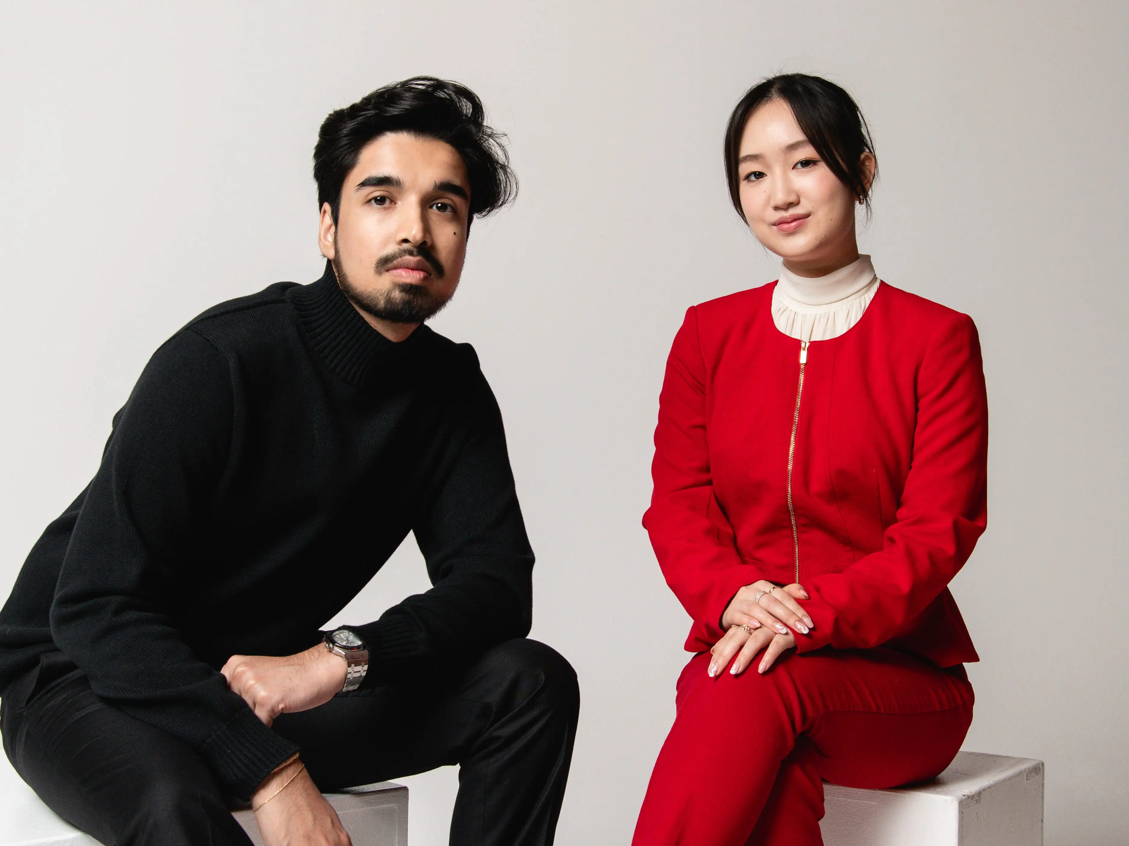 Clair Health cofounders Abhinav Agarwal and Jenny Duan, sitting on white cubes in front of a white background, wearing all black and all red -- respectively.