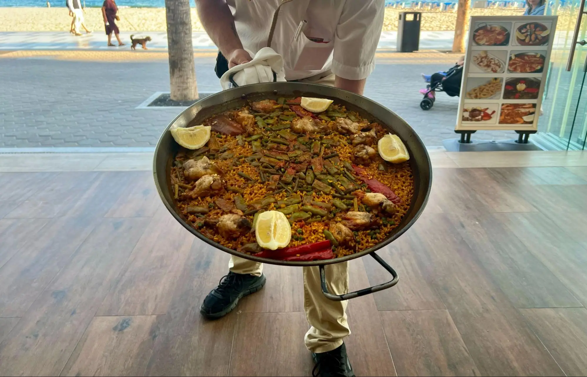 Restaurant server holds a large pan of paella with lemon wedges near a beachside entrance.