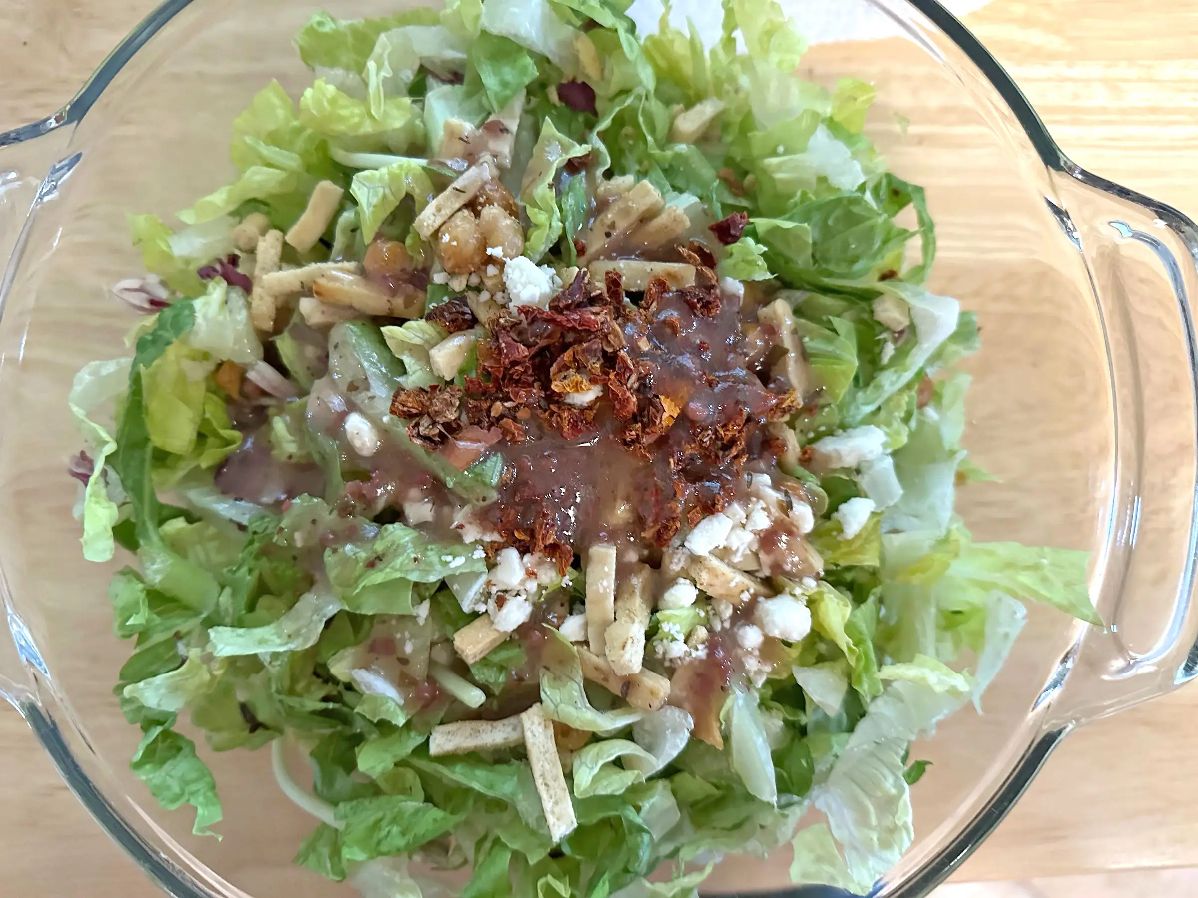 Green salad topped with dried tomato pieces, small flatbread sticks, and feta crumbles in a glass bowl.