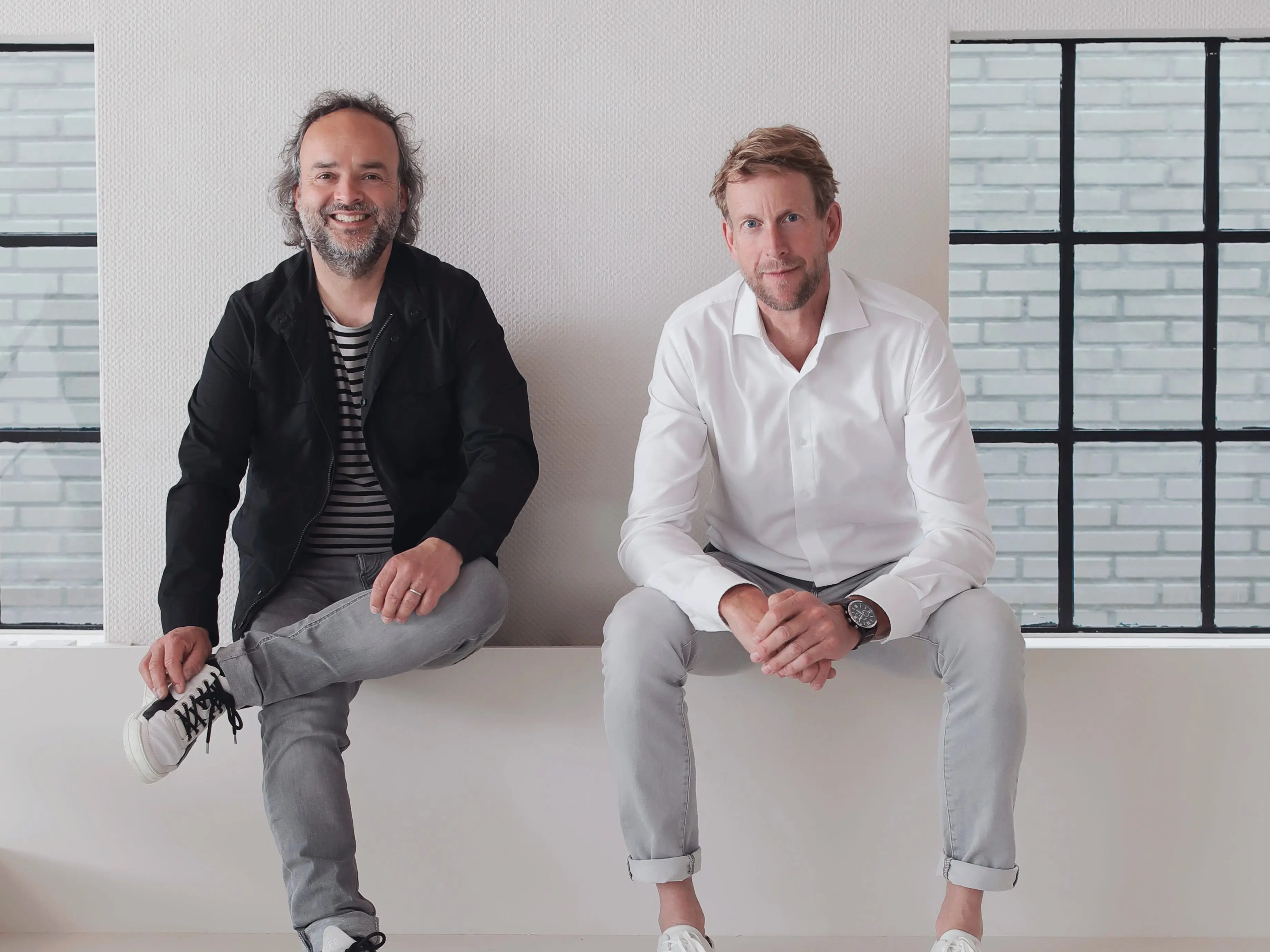 Two people sit on a white ledge above a sculptural table in a minimalist interior with large windows.
