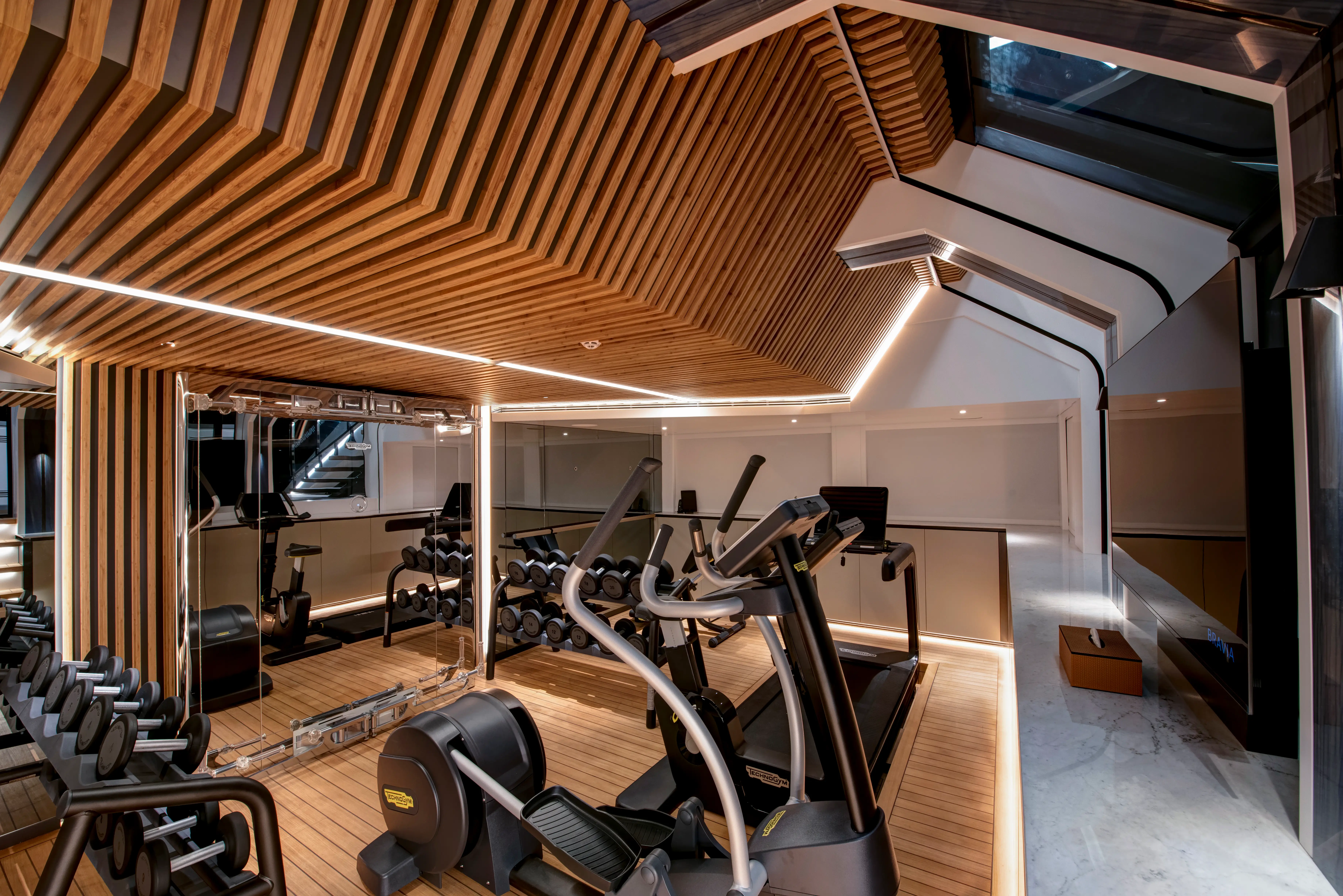 Modern fitness room with cardio machines, dumbbell racks, mirrors, and a slatted wood ceiling with linear lighting.