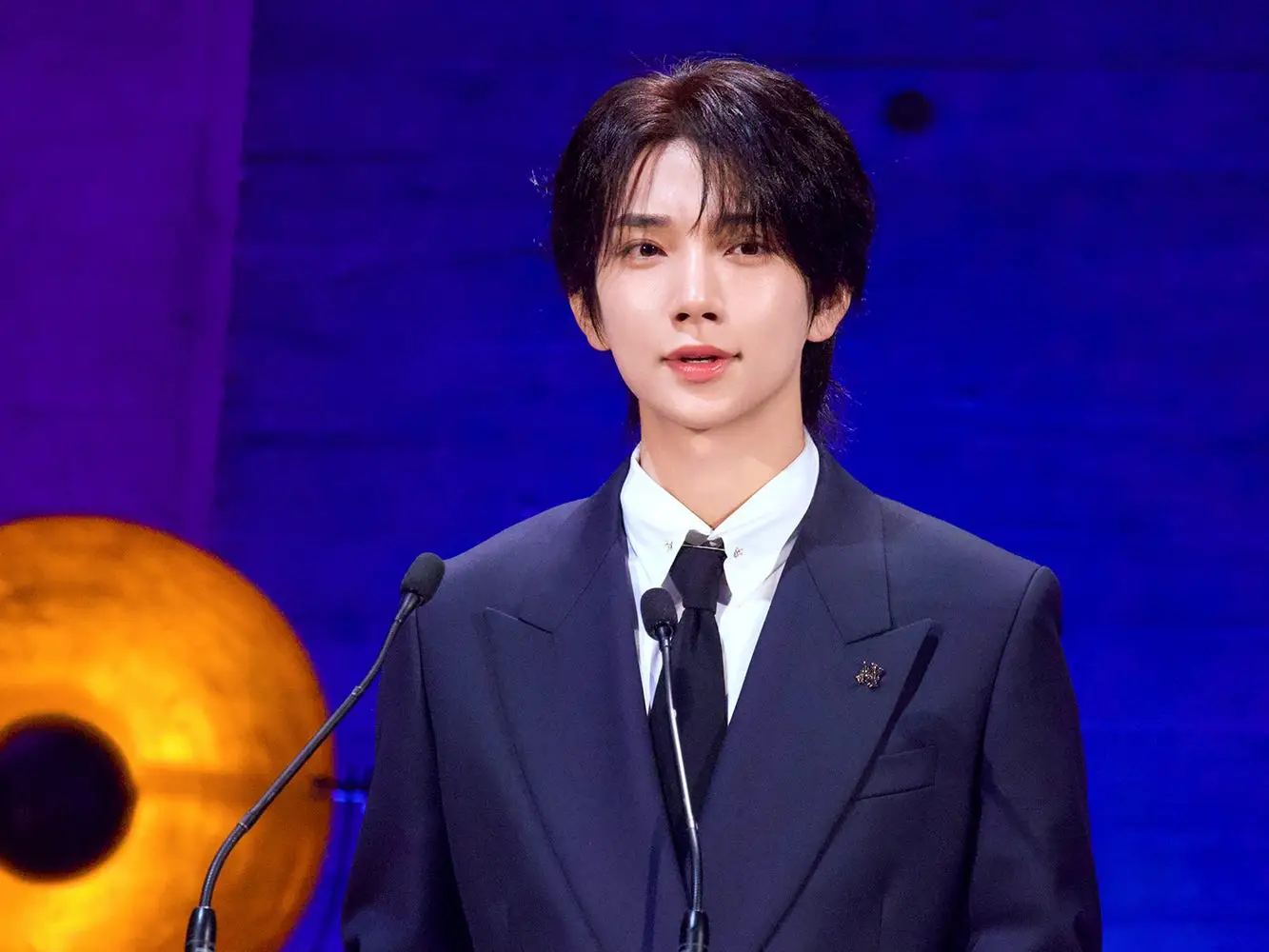 Joshua Hong, in a dark suit stands at a clear lectern with a blue unesco sign on a lit stage.
