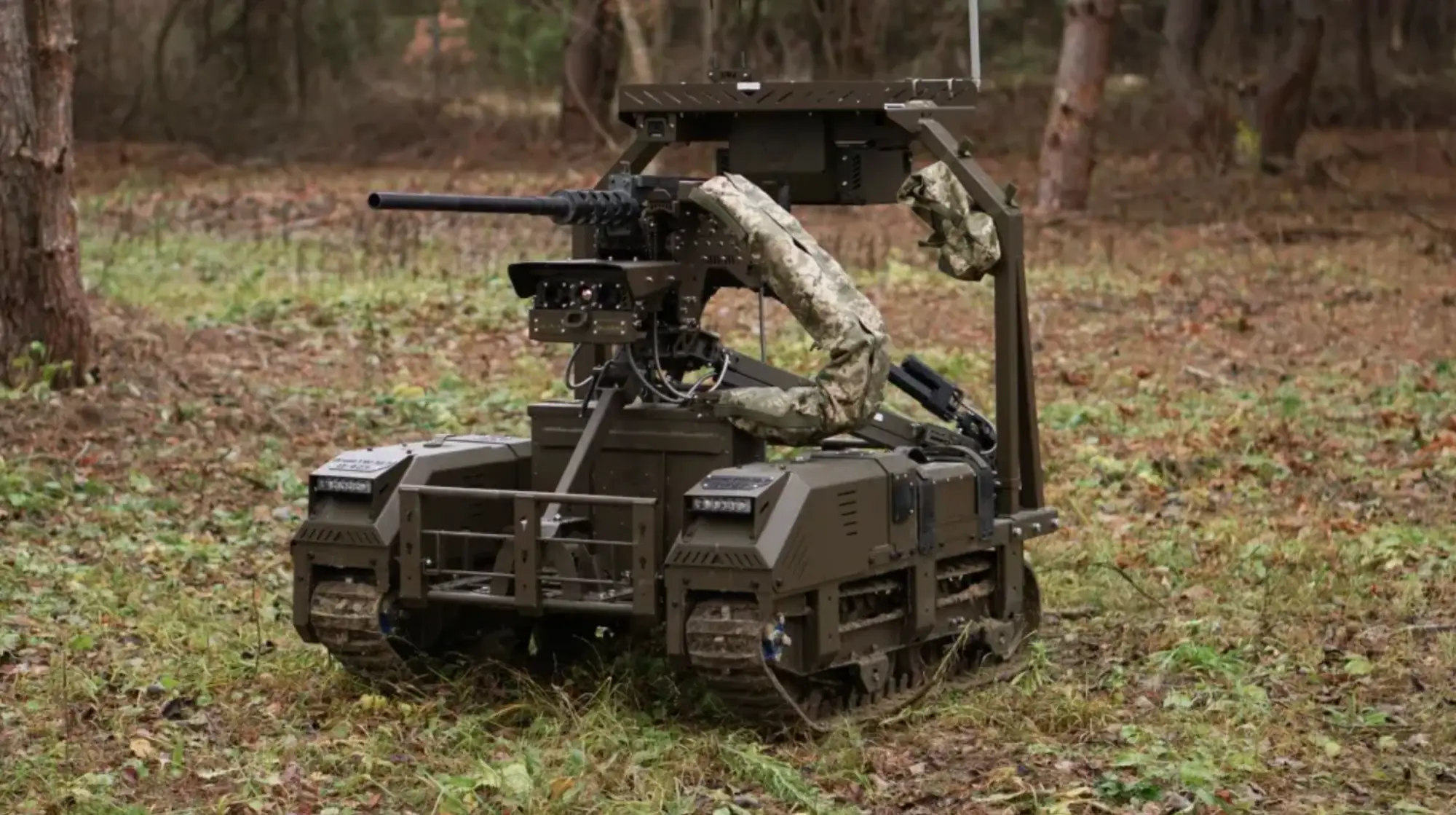 Tracked unmanned ground vehicle with a mounted weapon system positioned on grass in a wooded area.