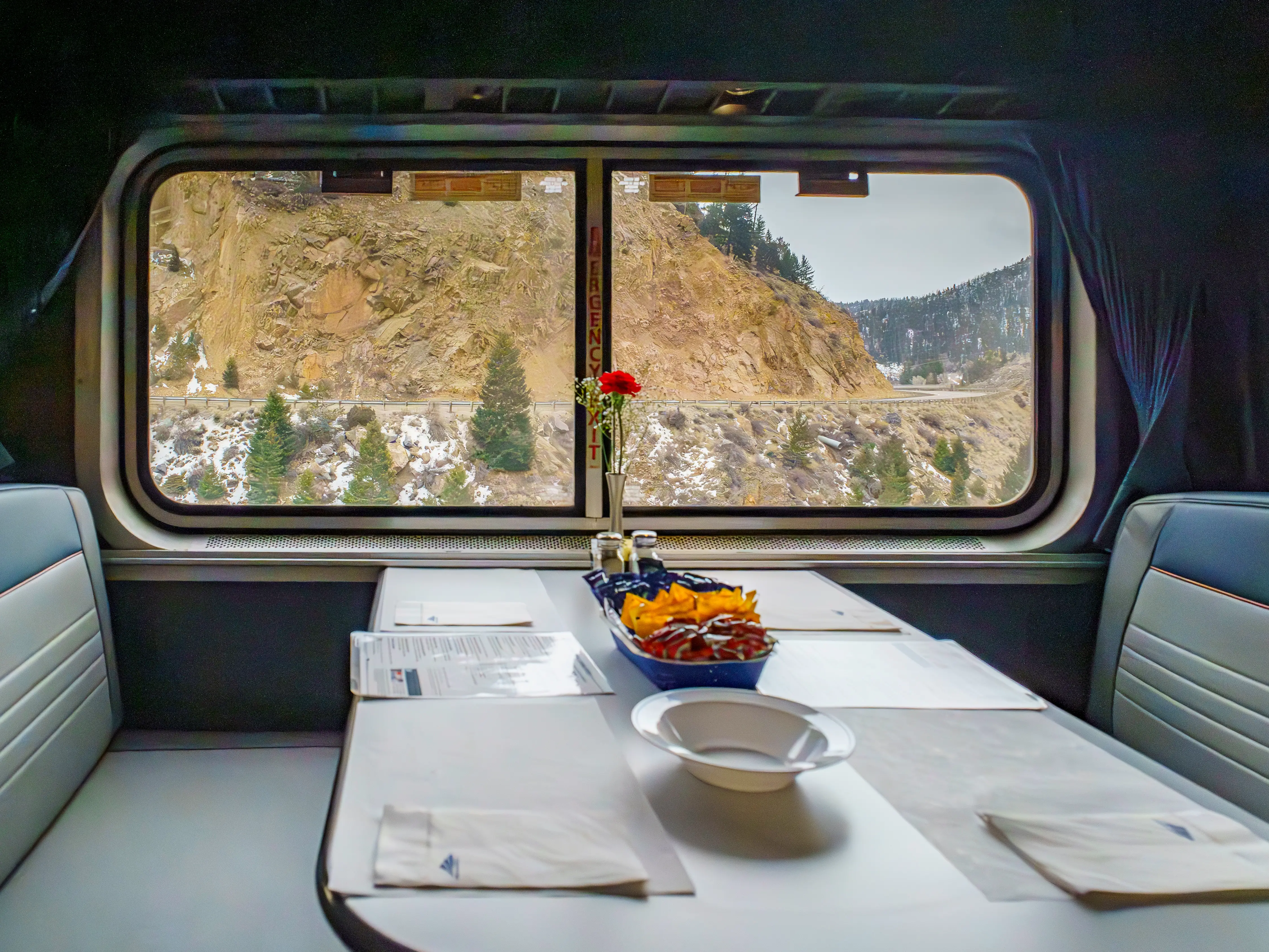 A booth in a train dining car with a wide window showing rocky mountains outside