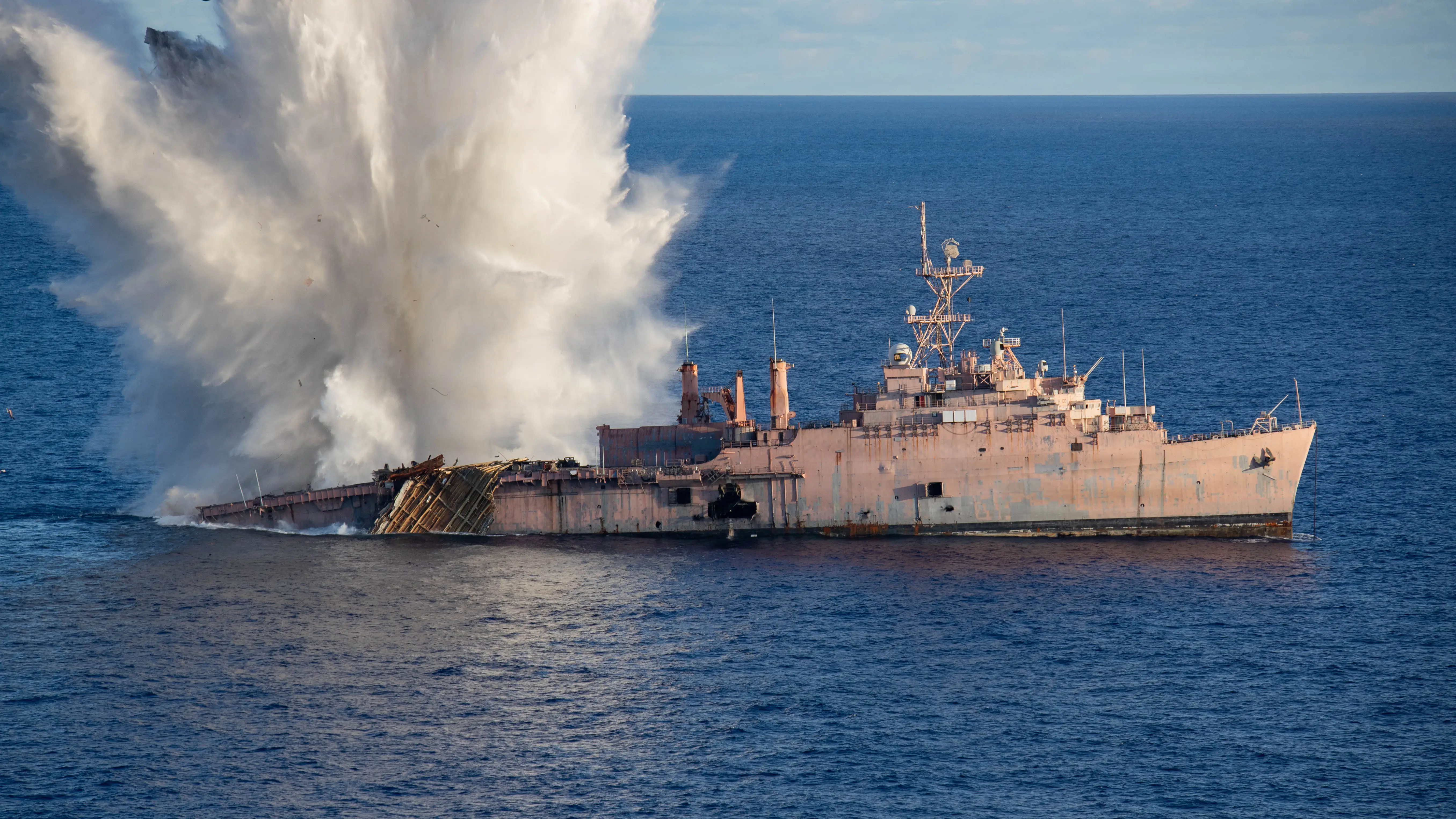 A large, decommissioned US Navy vessel is hit by a torpedo in dark blue ocean waters.