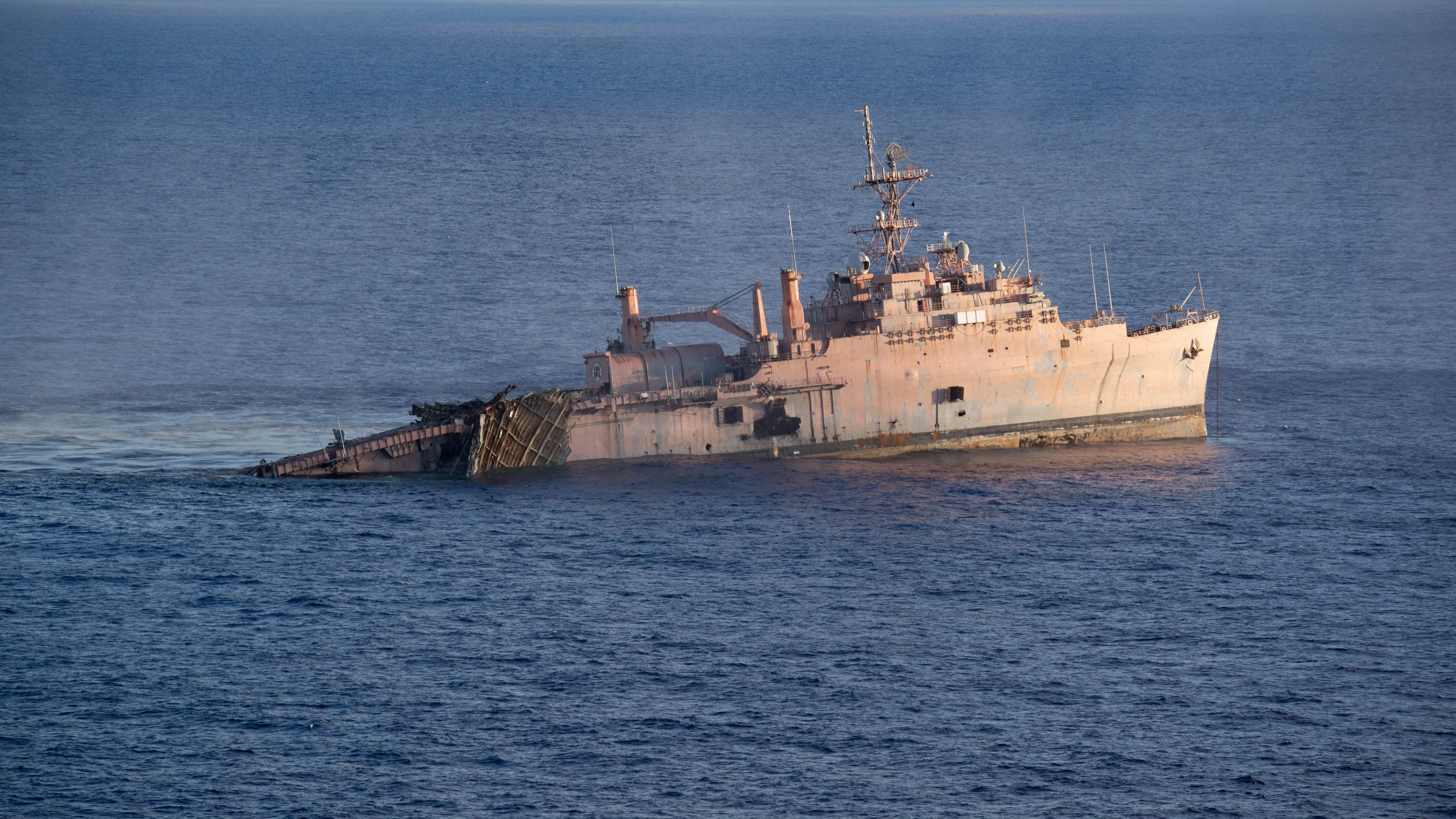 A large, decommissioned US Navy vessel starts to sink in dark blue ocean waters.