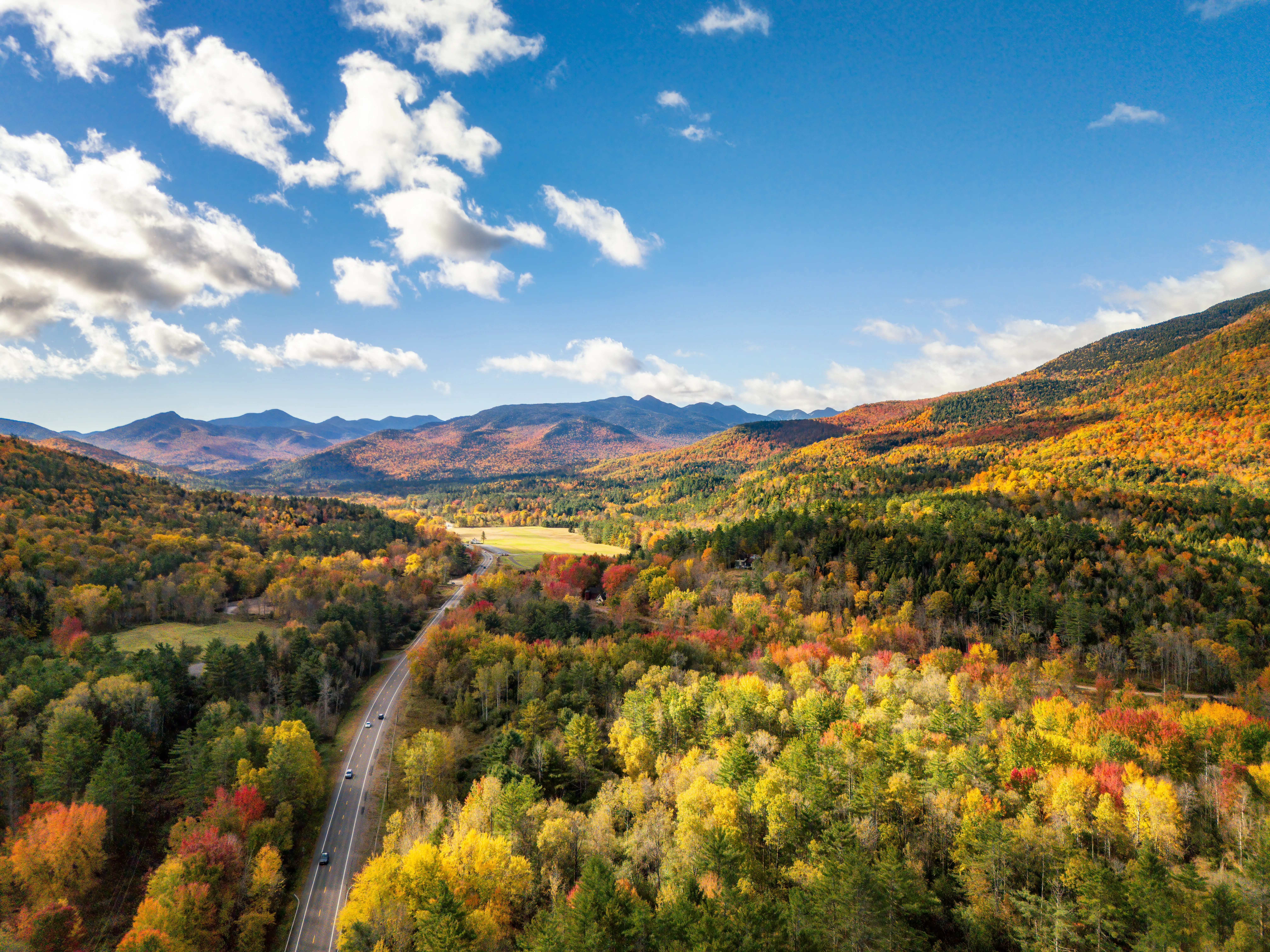 Aerial view of a winding road through colorful autumn forested mountains under a blue sky with scattered clouds.