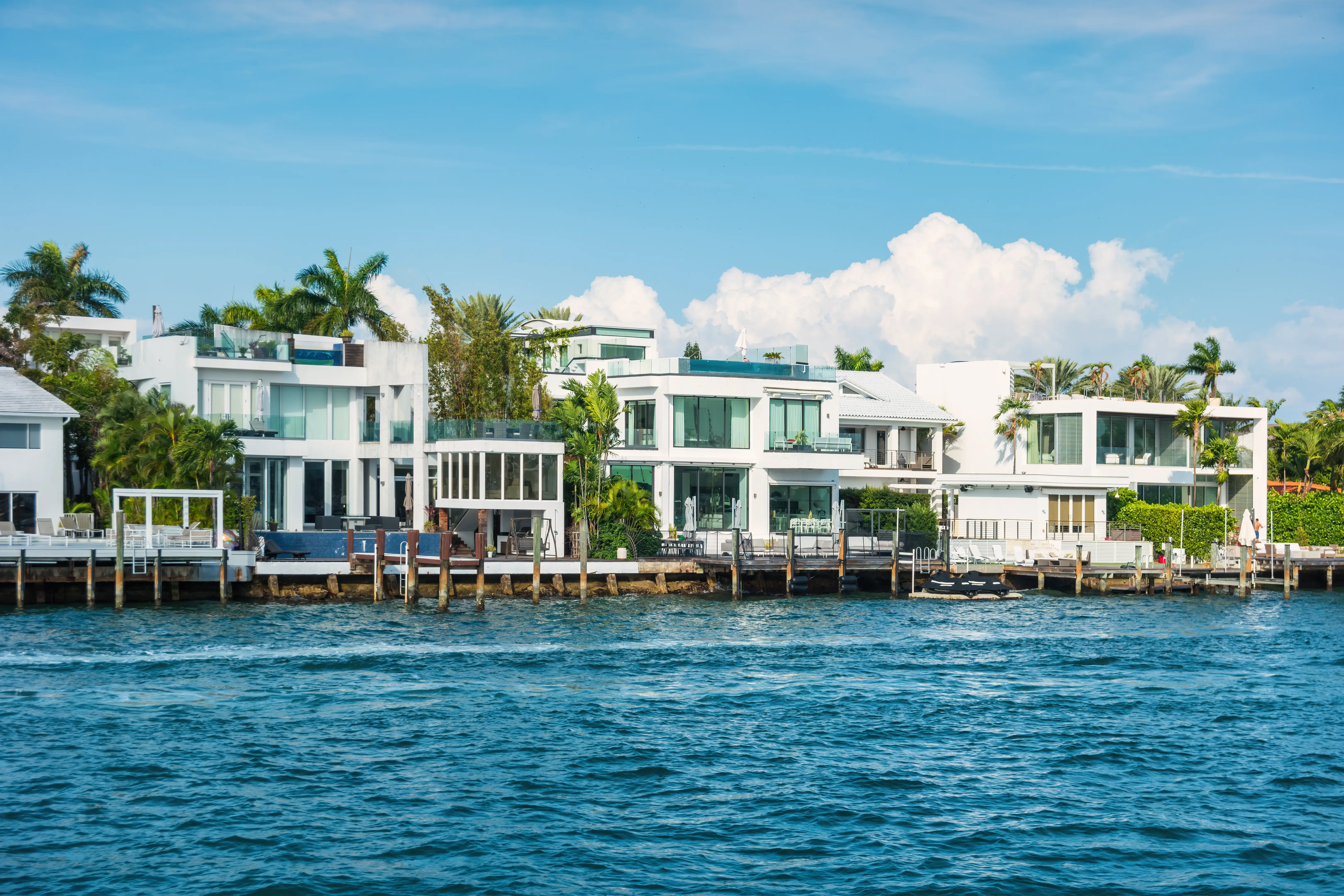 Modern waterfront homes with docks and palm trees line a bright blue waterway under a partly cloudy sky.