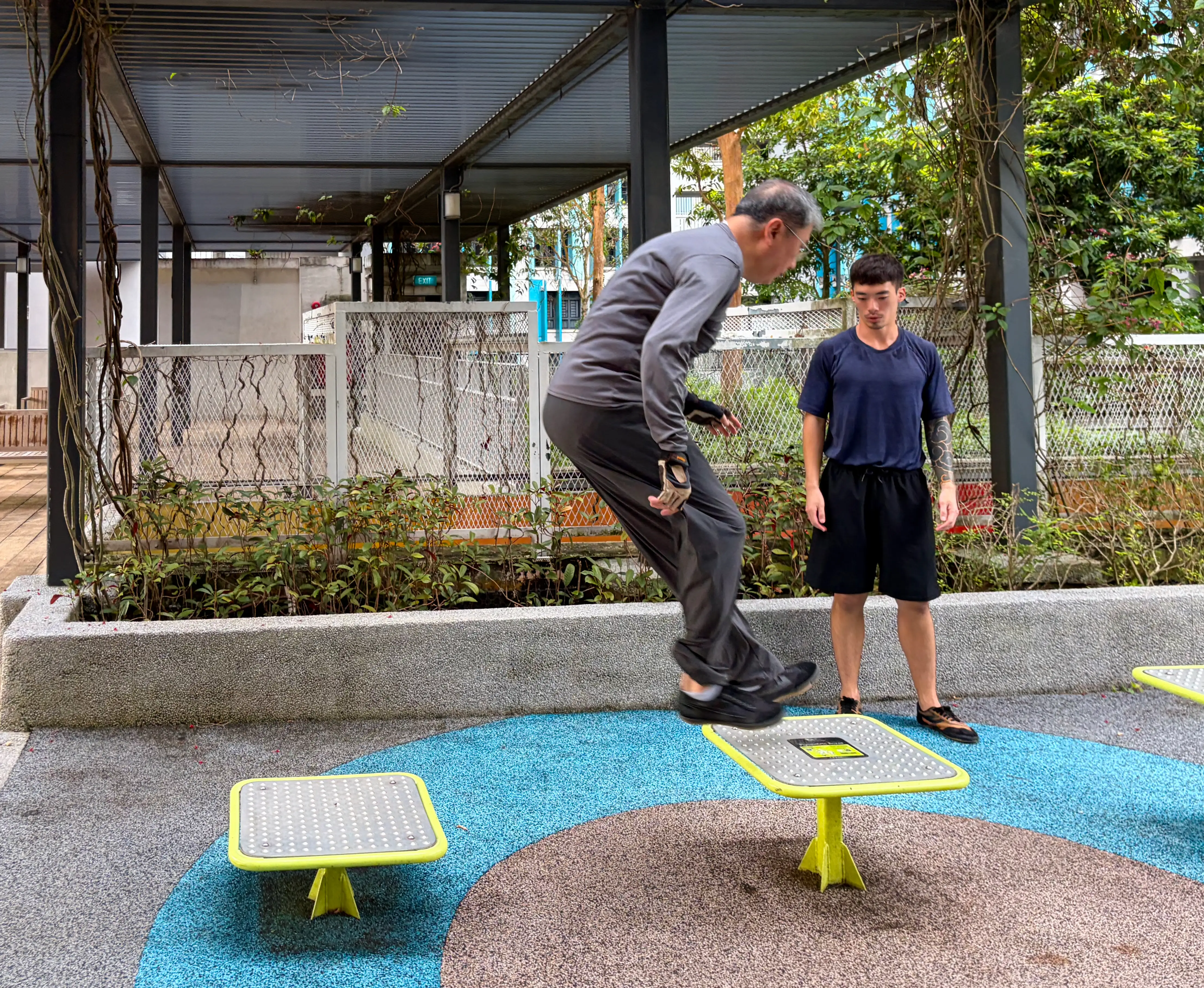A man watching as another older man does a jump across two playground structures.