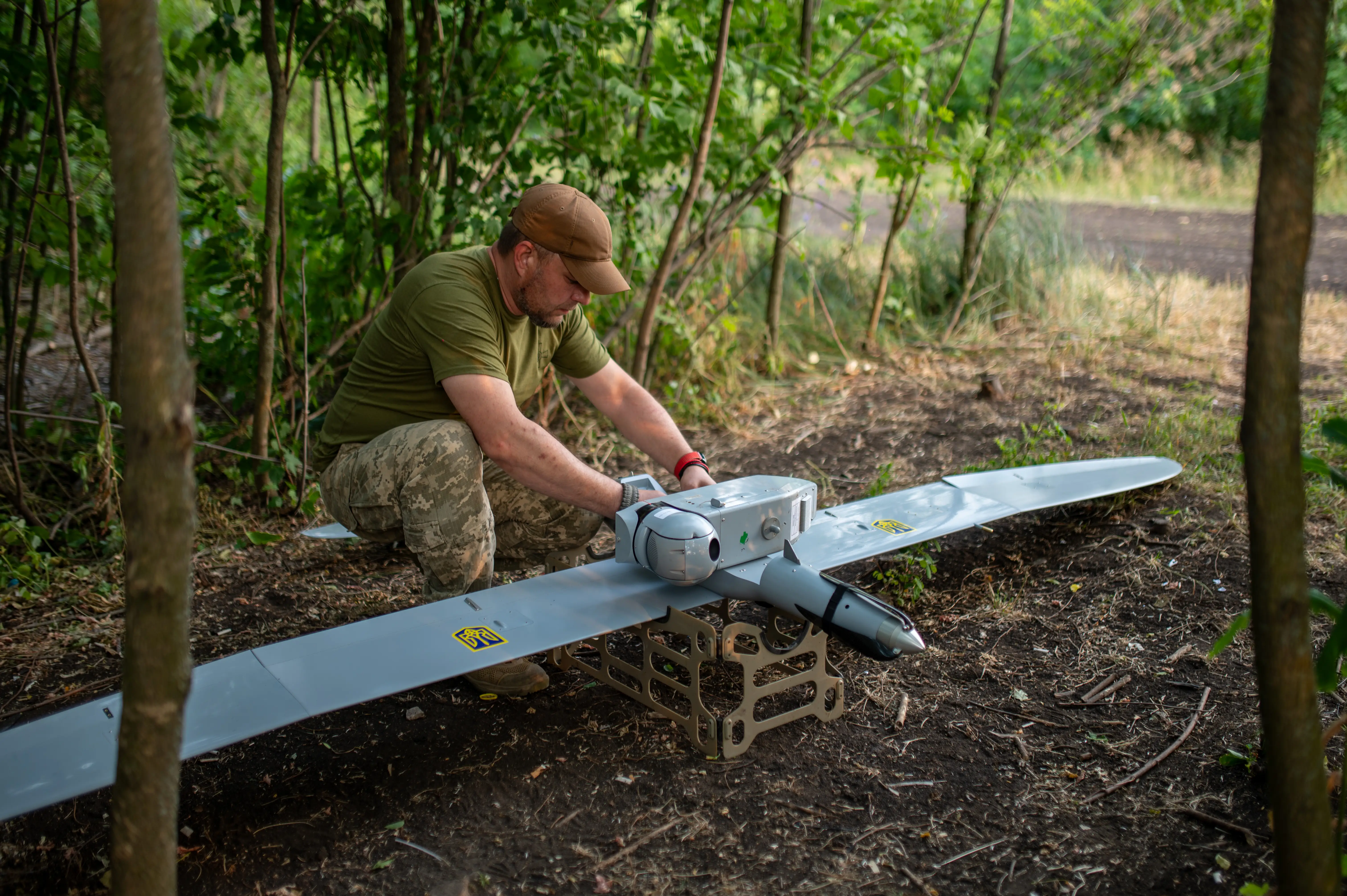 A man in khaki and a cap squats over a large grey drone among some trees