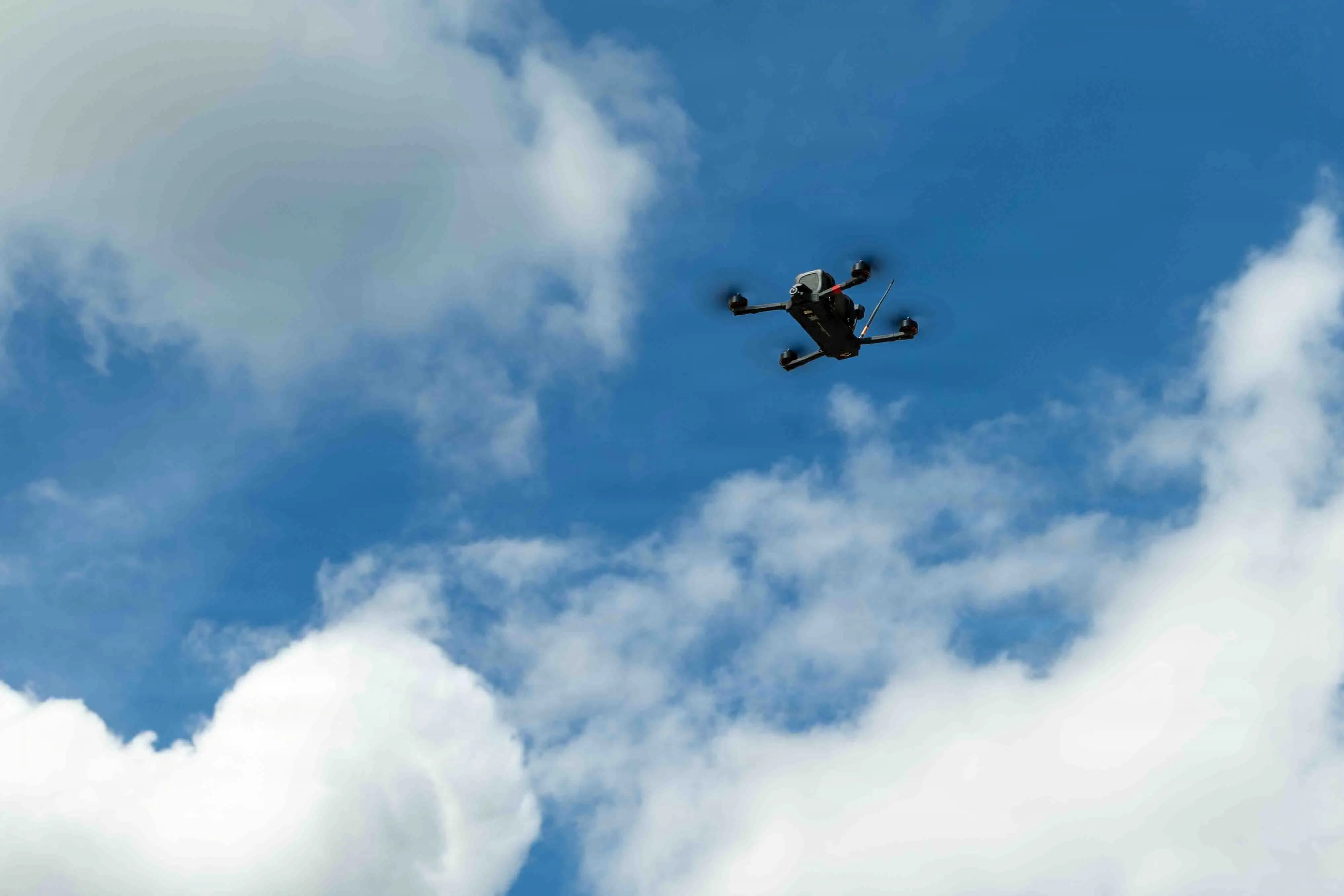 A small black drone flies in a cloudy sky.