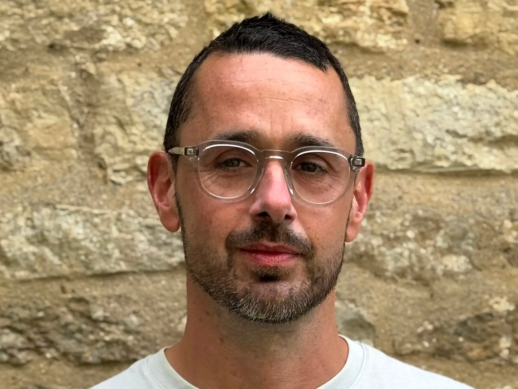 Portrait of a person wearing clear-frame glasses and a light shirt in front of a stone wall.