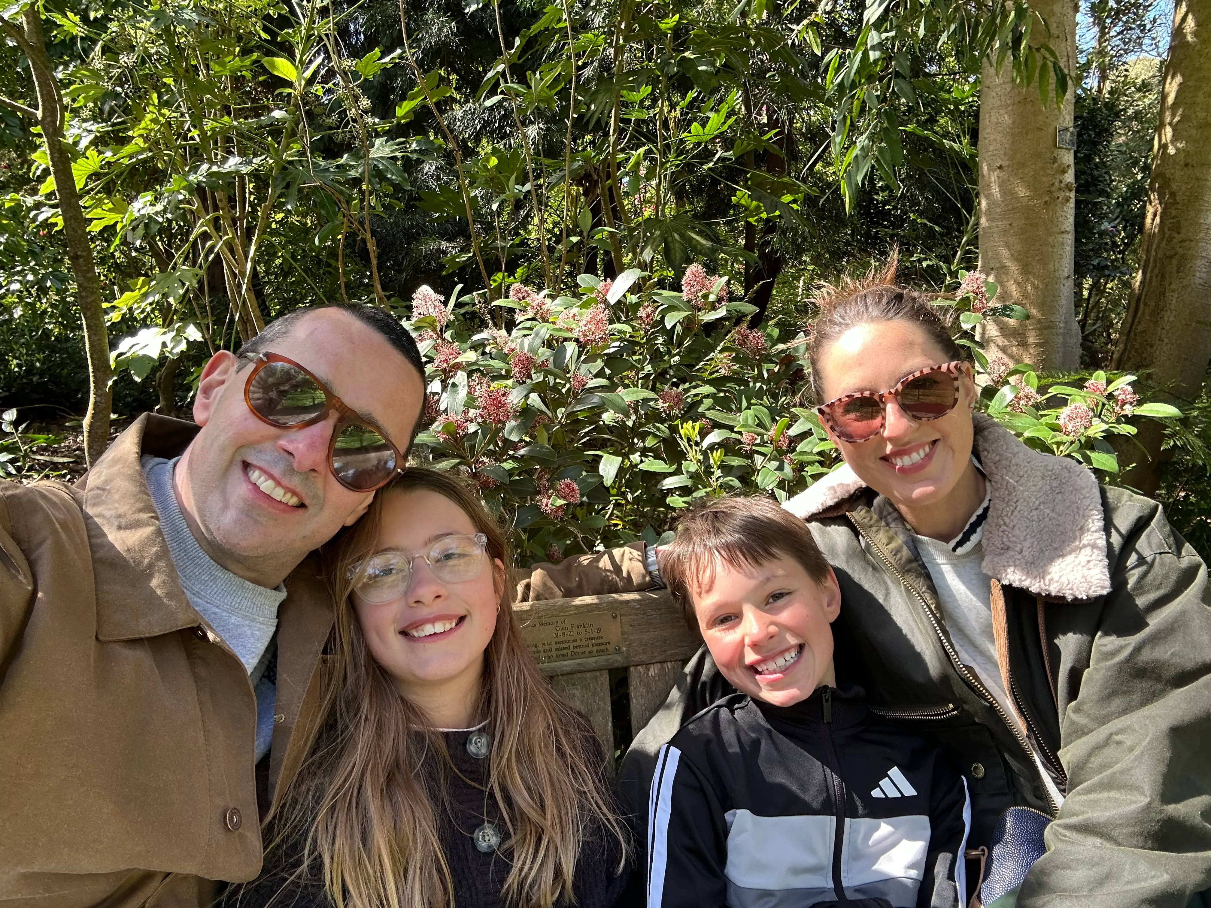 Four smiling people pose for a selfie on a wooden bench in a sunny garden with flowering shrubs.