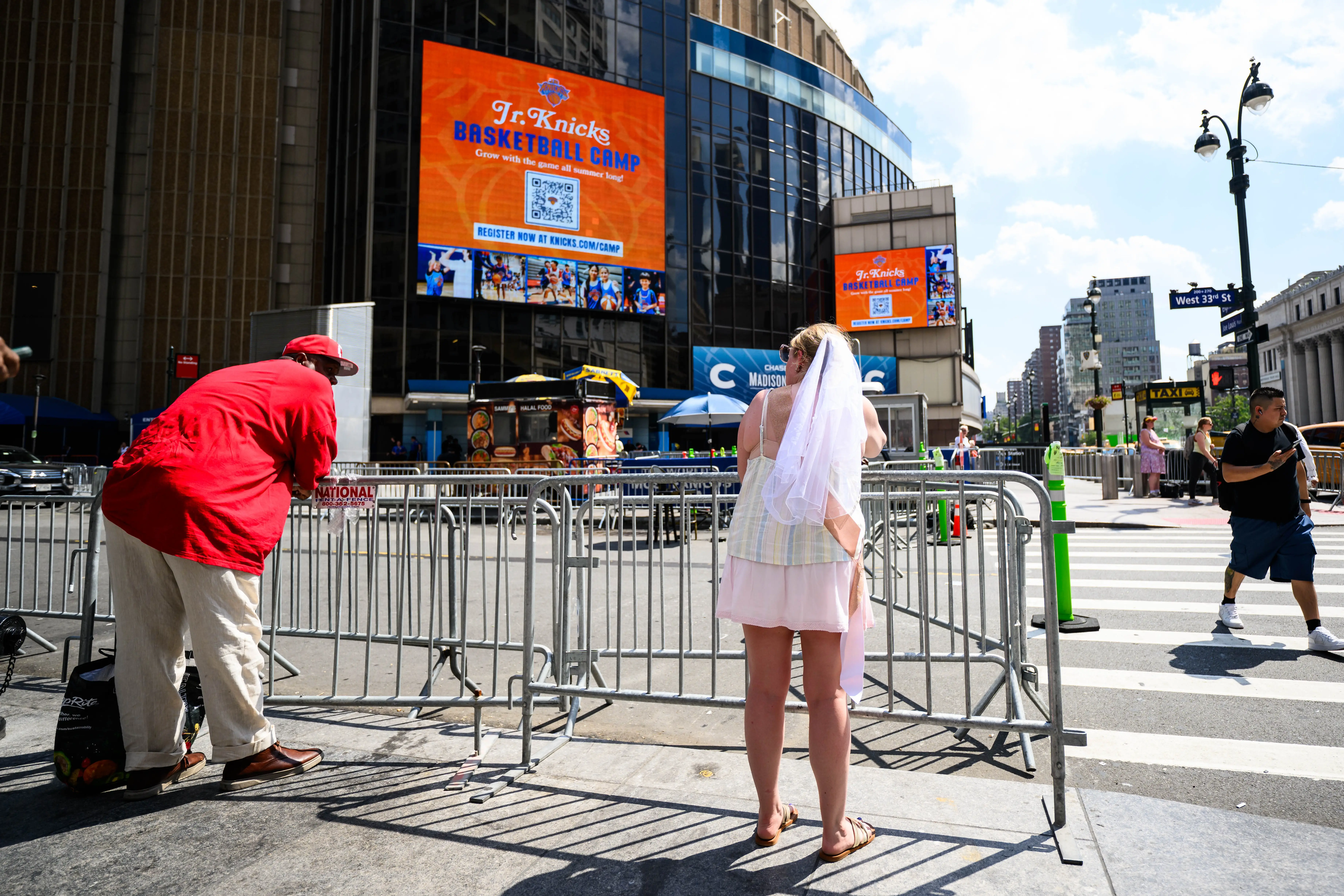 A woman wearing a veil wedding veil land a man in a red jacket lean on a police barricade and look up and down the street outside Madison Square Garden