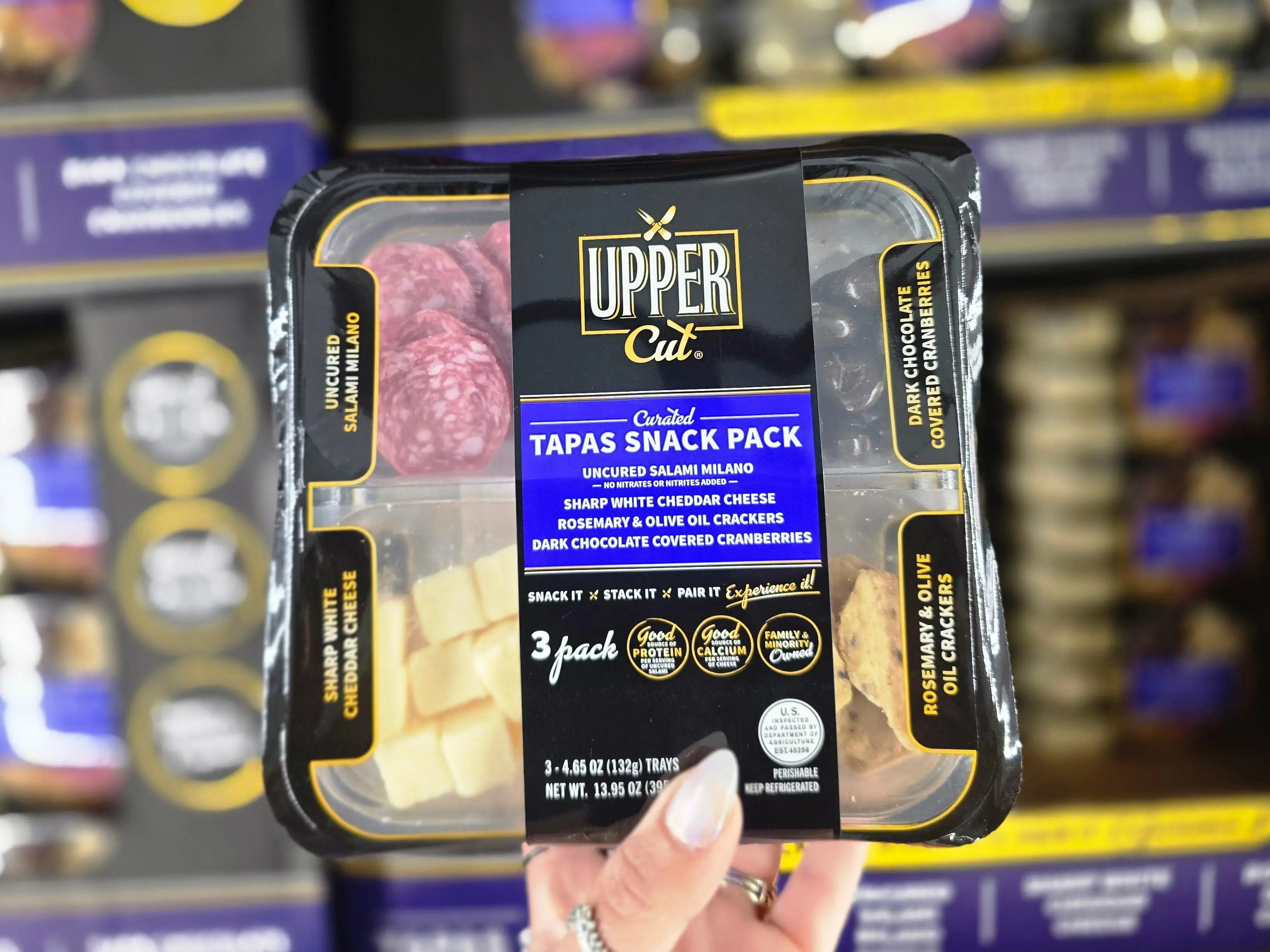 A hand holding an Upper Cut snack pack with salami, cheese, crackers, and chocolate-covered cranberries.