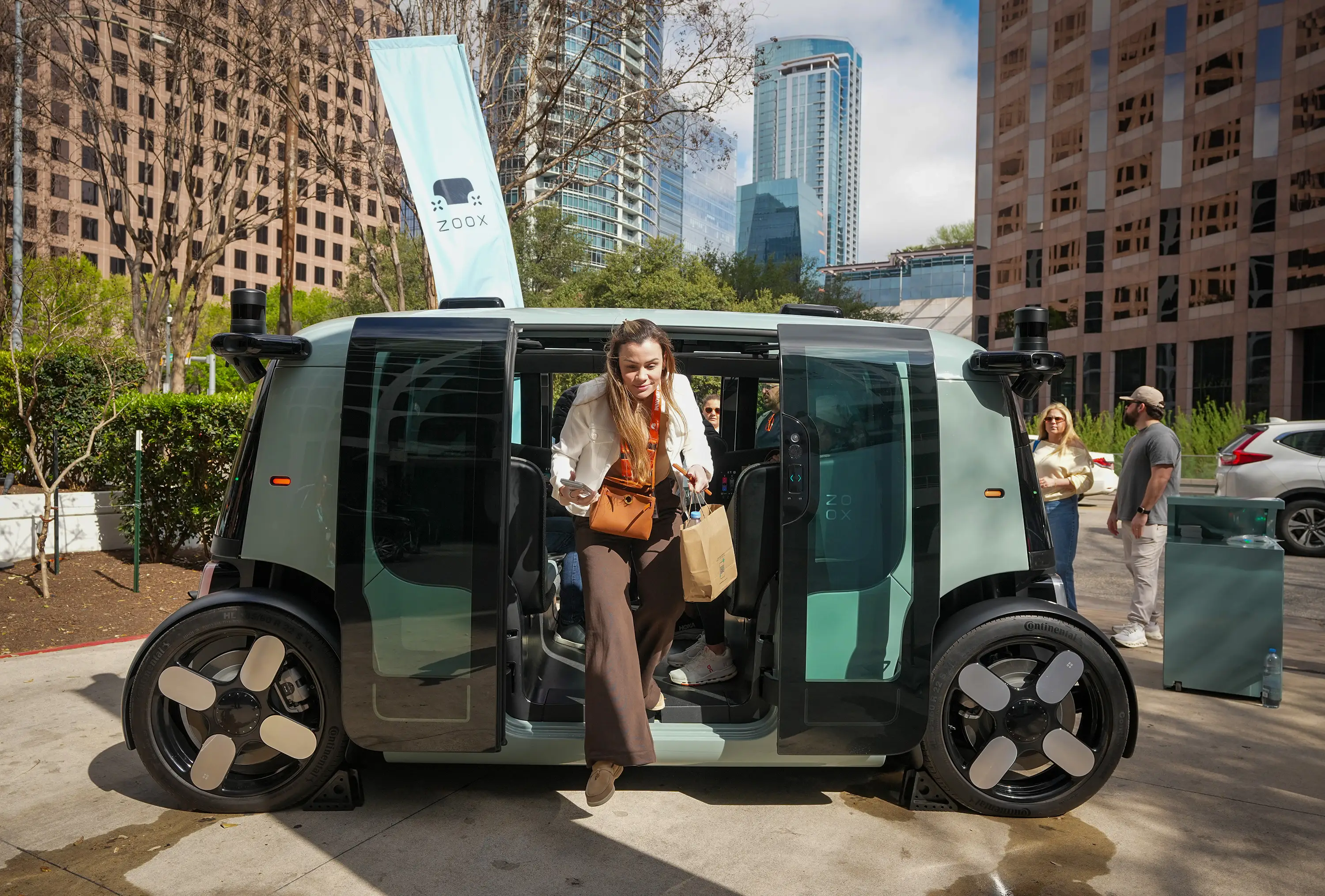 A woman carrying a brown paper shopping bag steps out of a Zoox self-driving robotaxi as a light green Zoox banner sits behind it and skyscrapers stand in the distance.