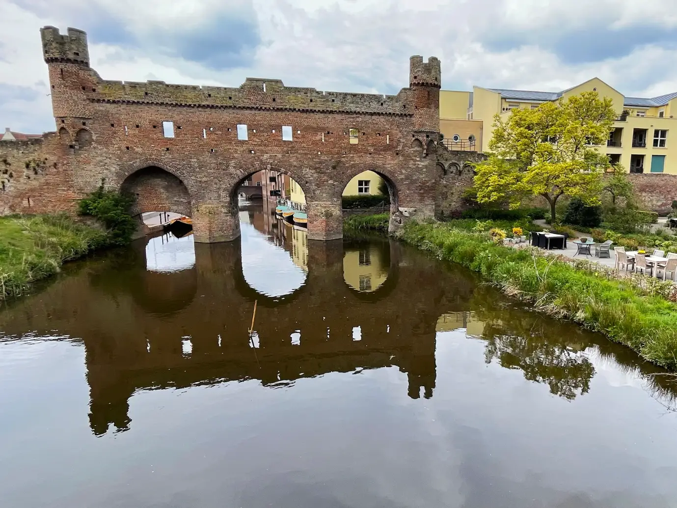 A medieval brick gate spans a calm canal in the Netherlands with buildings and garden seating nearby.