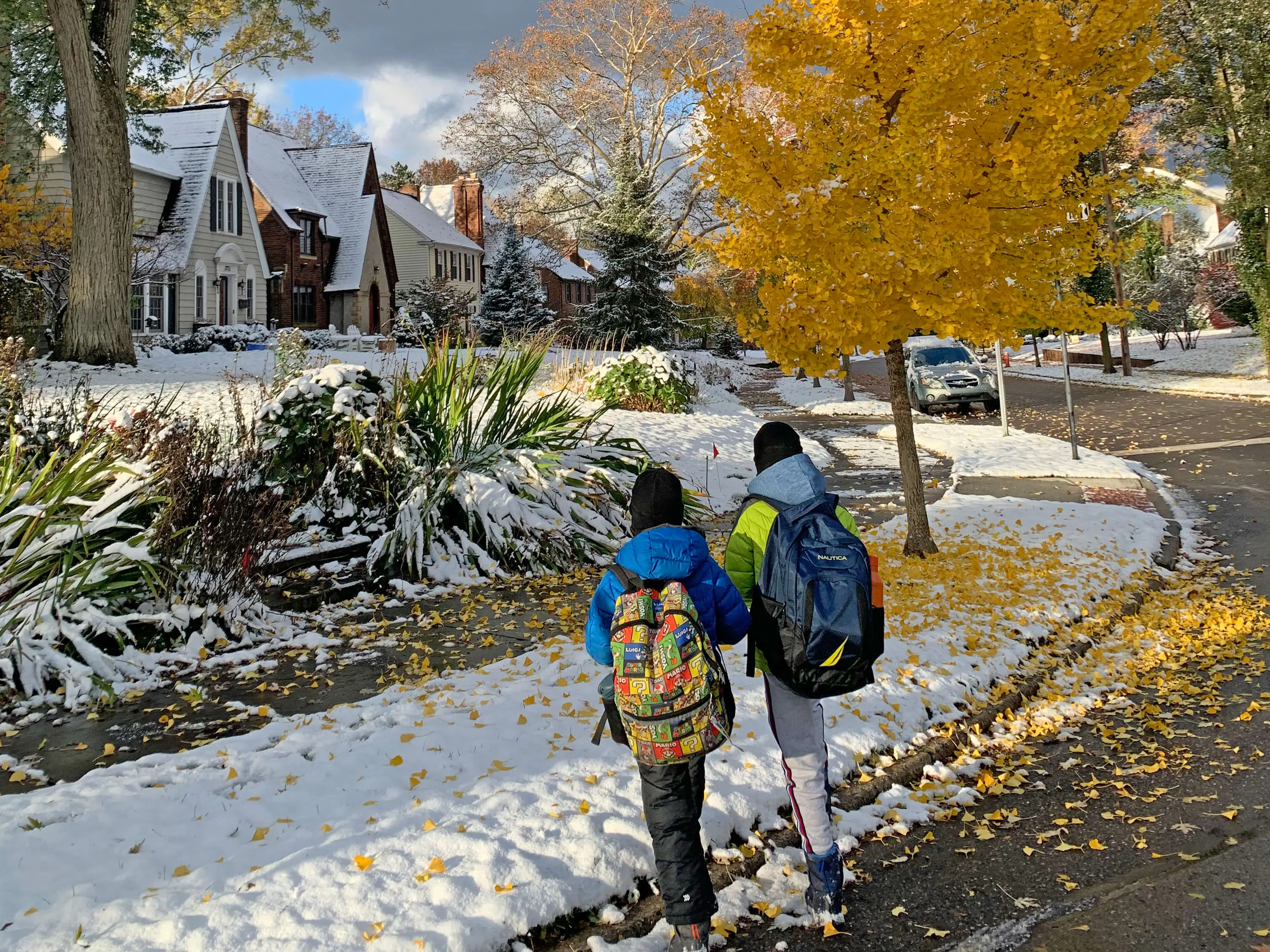 Two kids walking with backpacks on a fall day with snow on the ground.