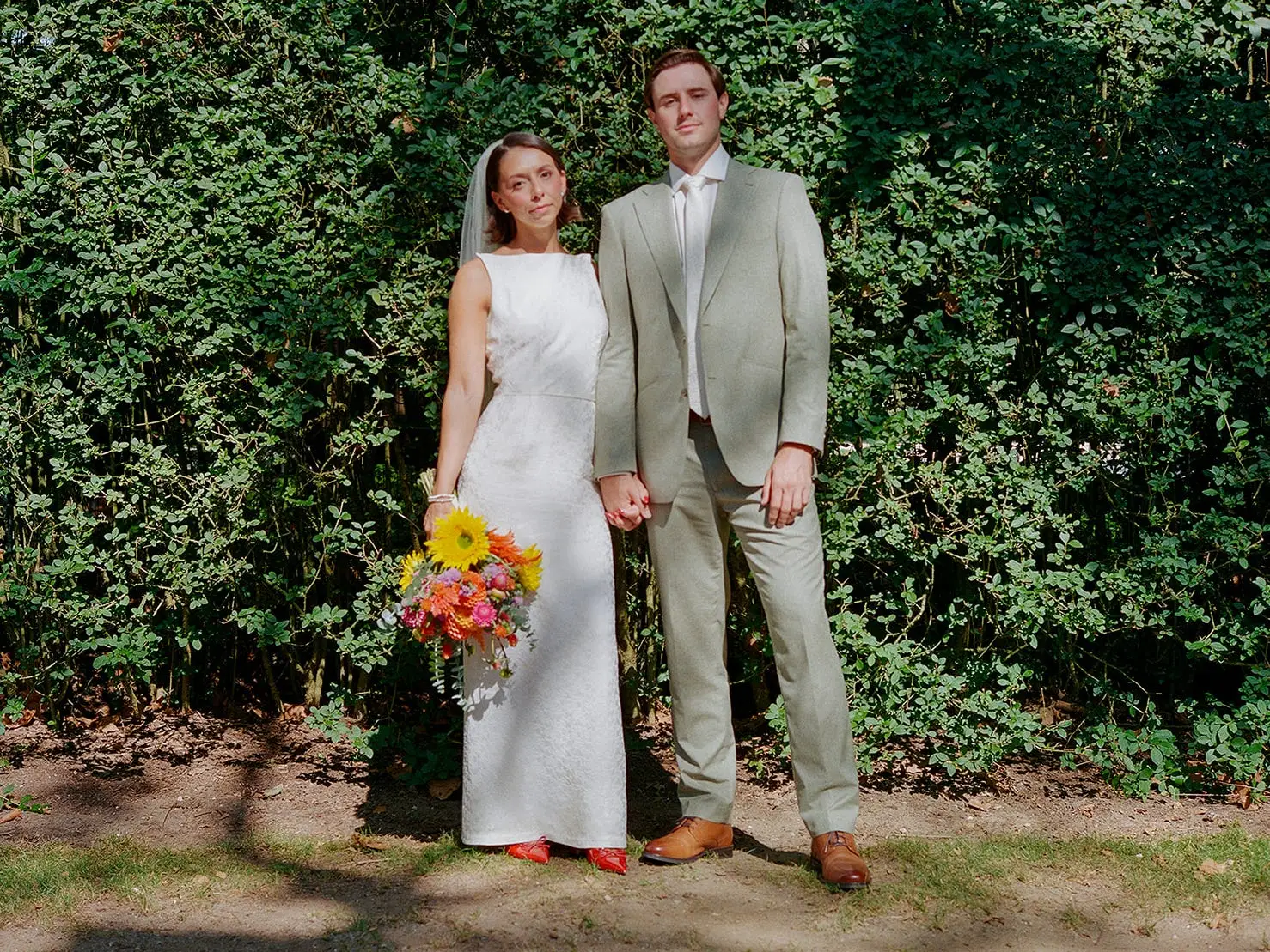 A bride and groom stand in front of a wall of greenery.