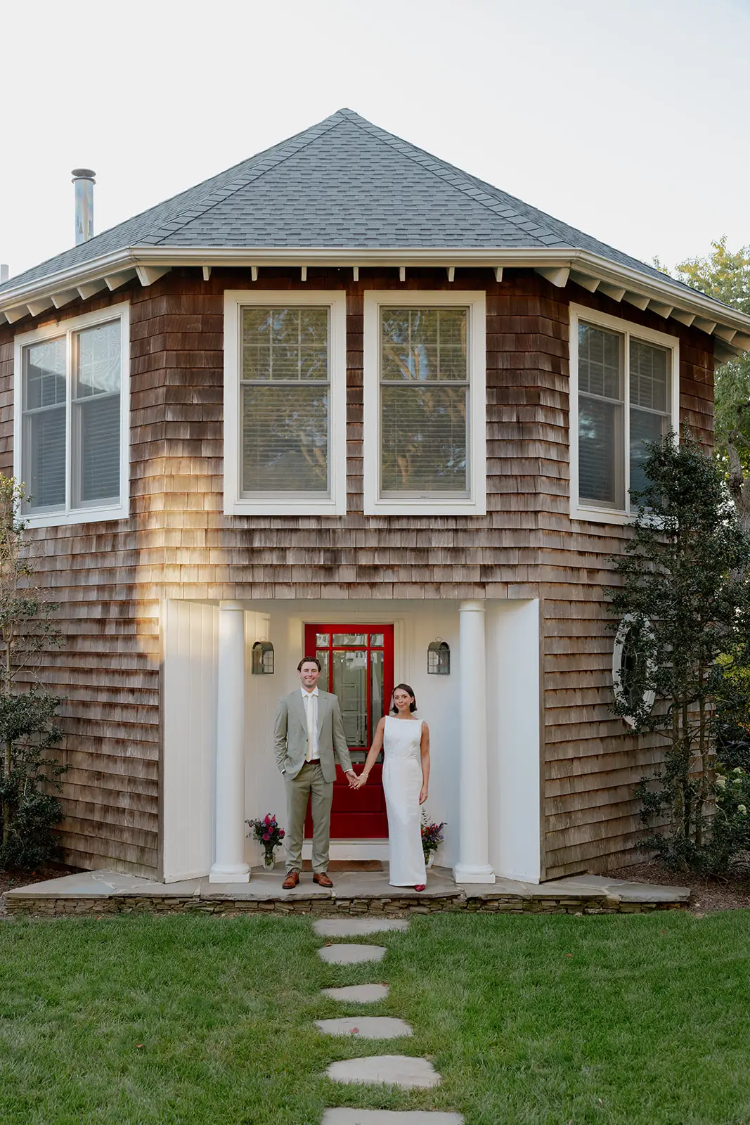 A bride and groom stands in front of a rounded house with a red door.