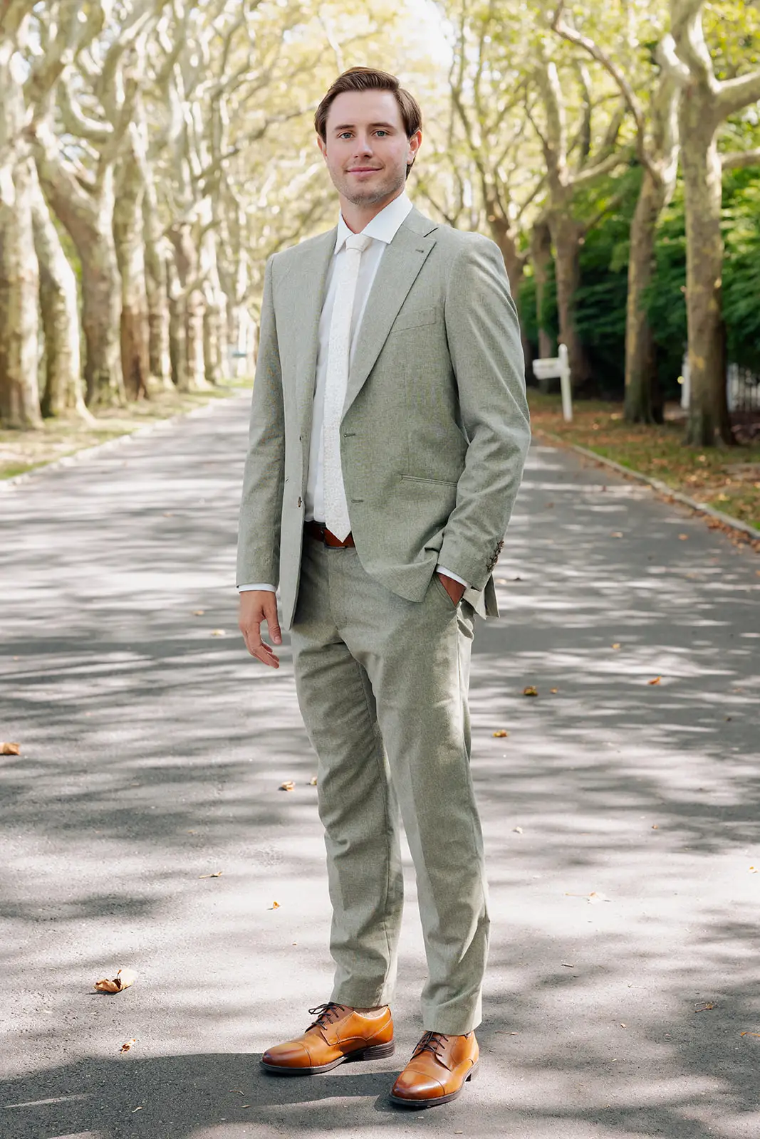A groom stands in a green suit on a tree-lined street.