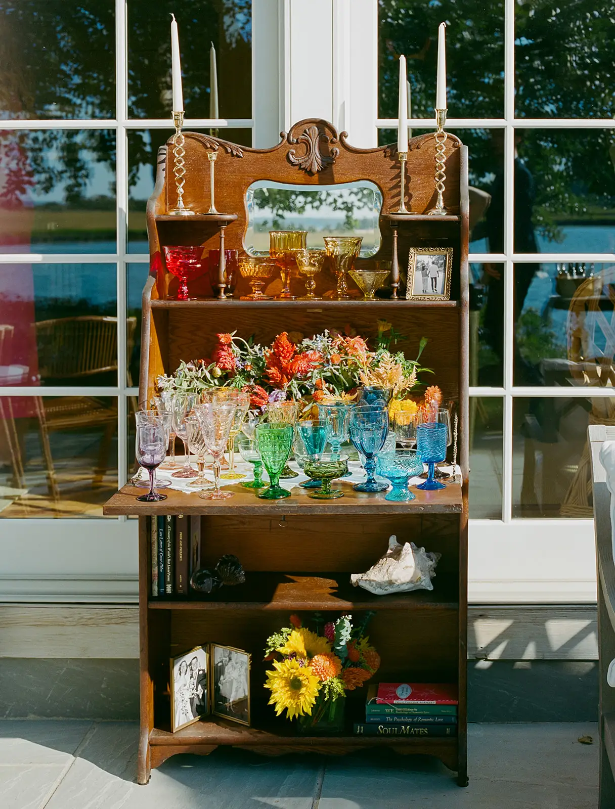 A wooden hutch with colorful glasses, candles, and flowers displayed.
