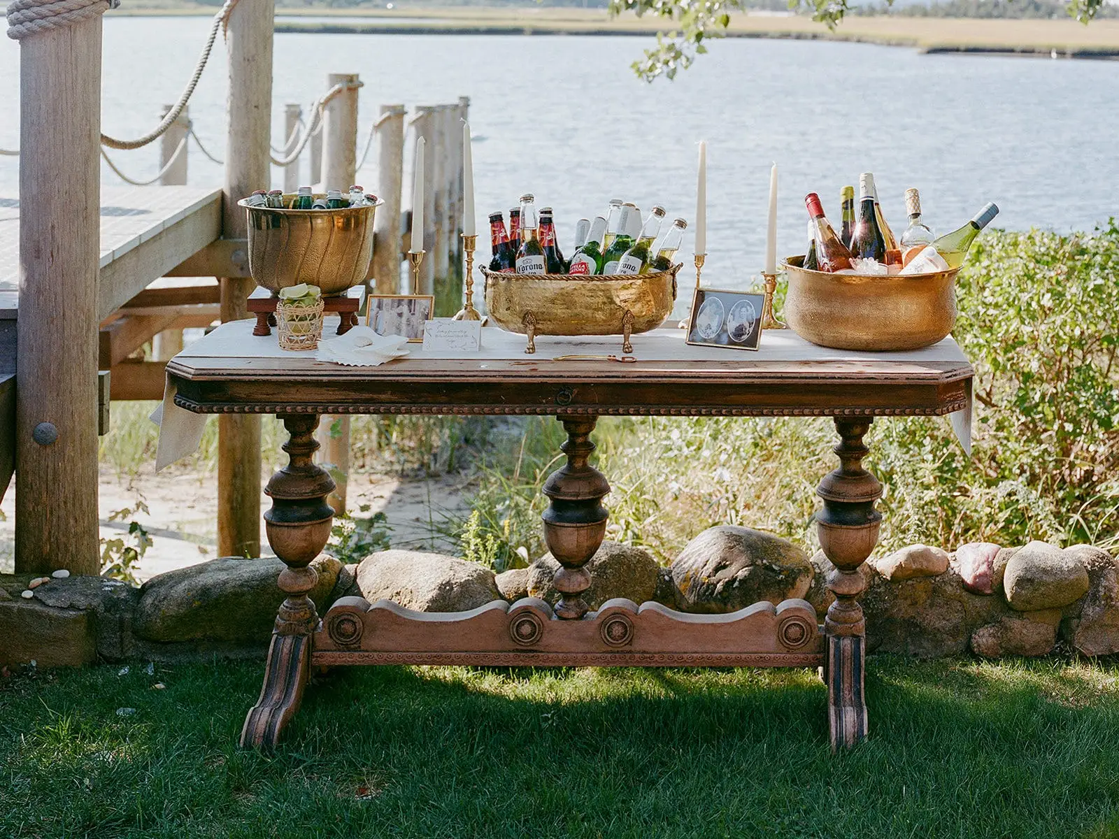 A wooden table with gold bowls full of alcohol on it. Flowers and candles sit ont he table as well, which sits in front of a body of water.