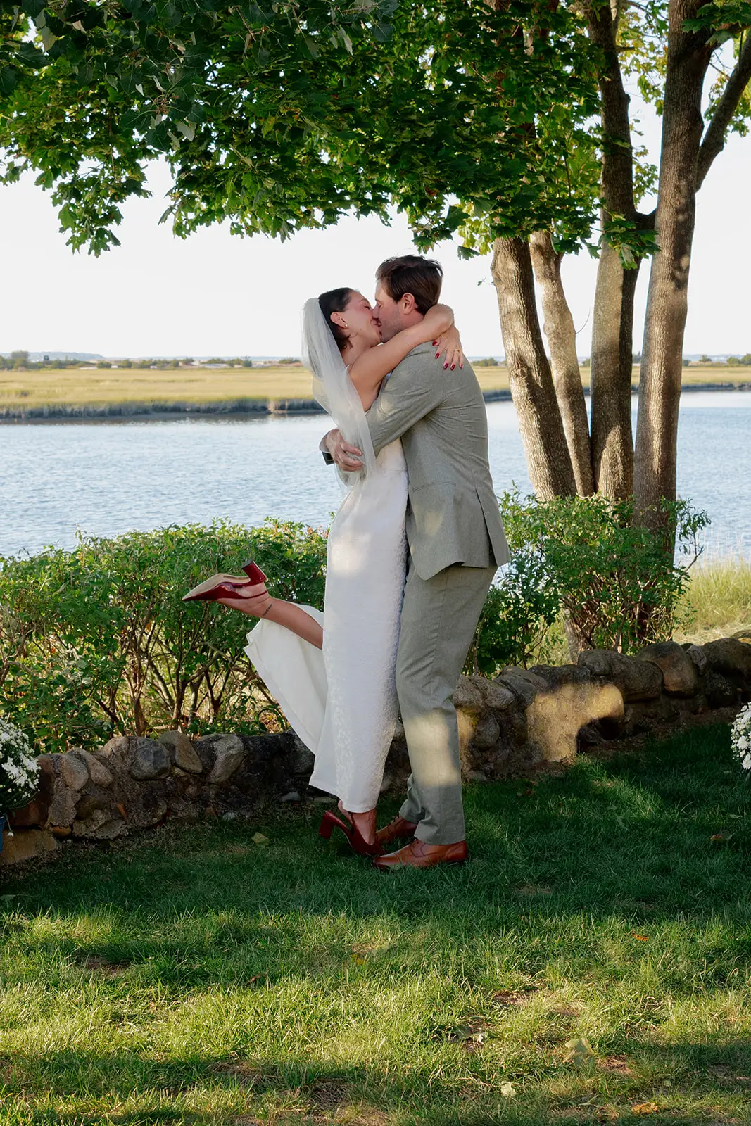 A bride and groom kiss in front of water. The groom lifts the bride off her feet and she kicks a leg up.
