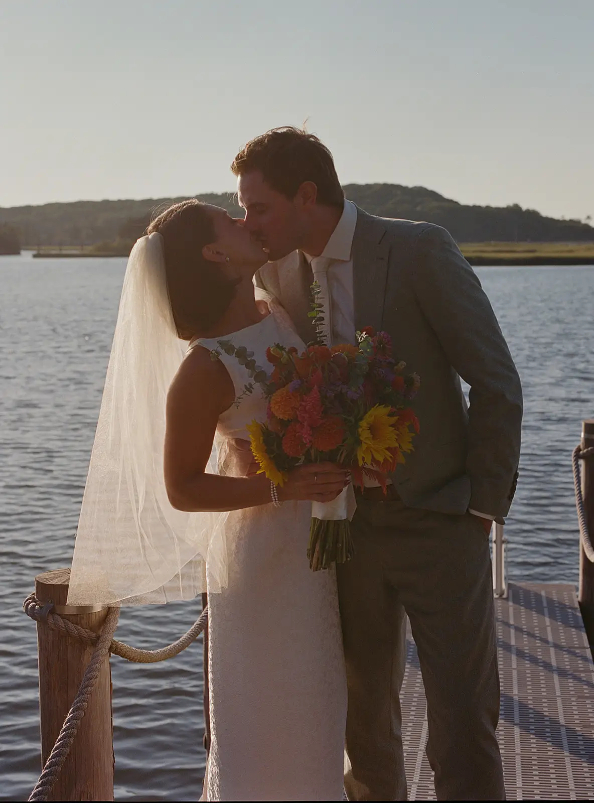 A bride and groom kiss in front of a body of water on a dock.