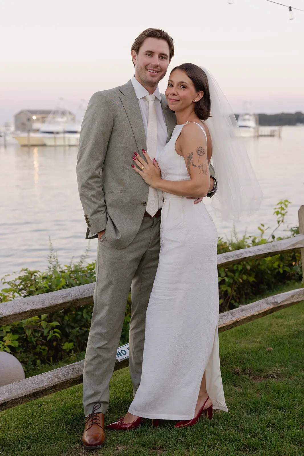 A bride and groom stand in front of water on a grassy lawn.