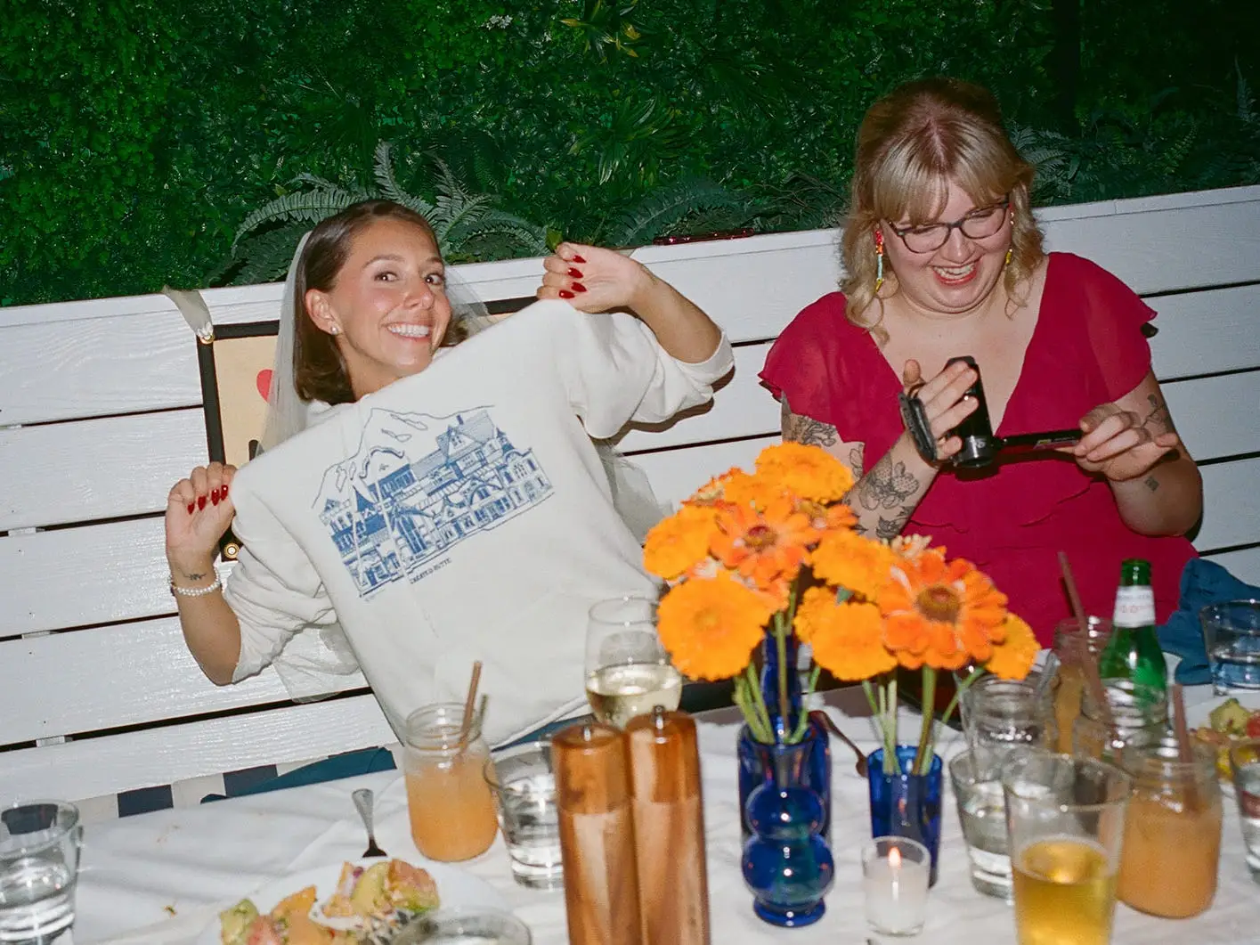 A bride holds up a sweatshirt she's wearing as a friend laughs next to her and holds up a camcorder.