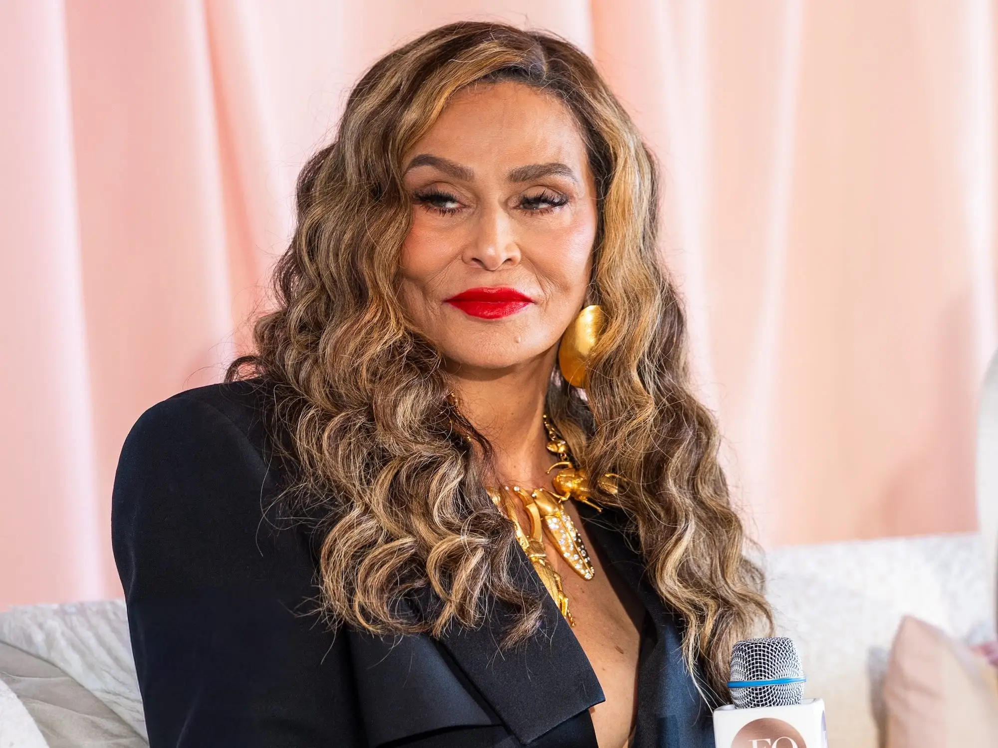 Tina Knowles sits on a white couch holding a microphone before a pink draped backdrop.